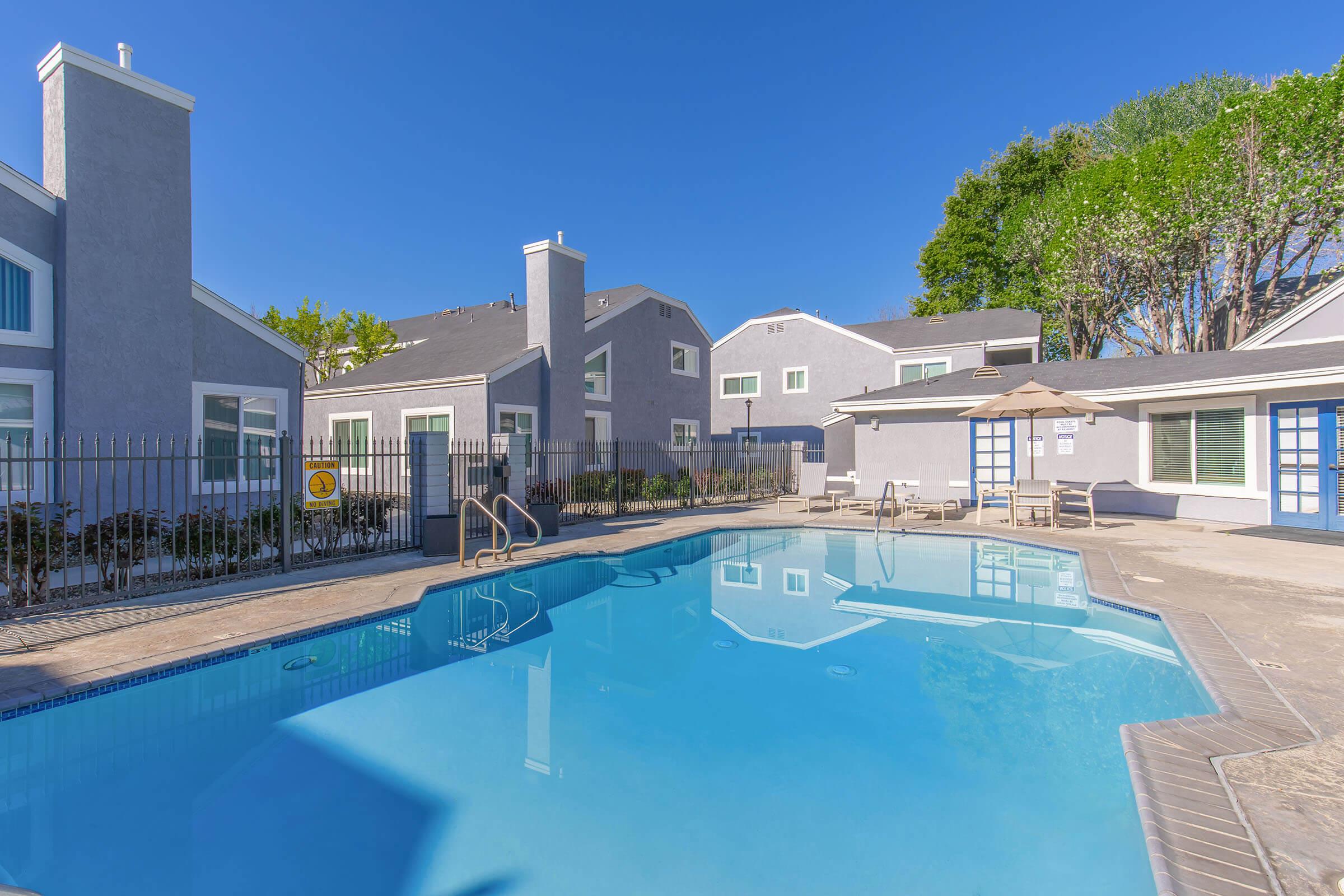 A clear blue swimming pool is situated in a sunny courtyard surrounded by residential buildings with gray exteriors. The pool area features lounge chairs and a shaded seating area, all enclosed by a black fence. Bright blue skies and well-maintained greenery are visible in the background.