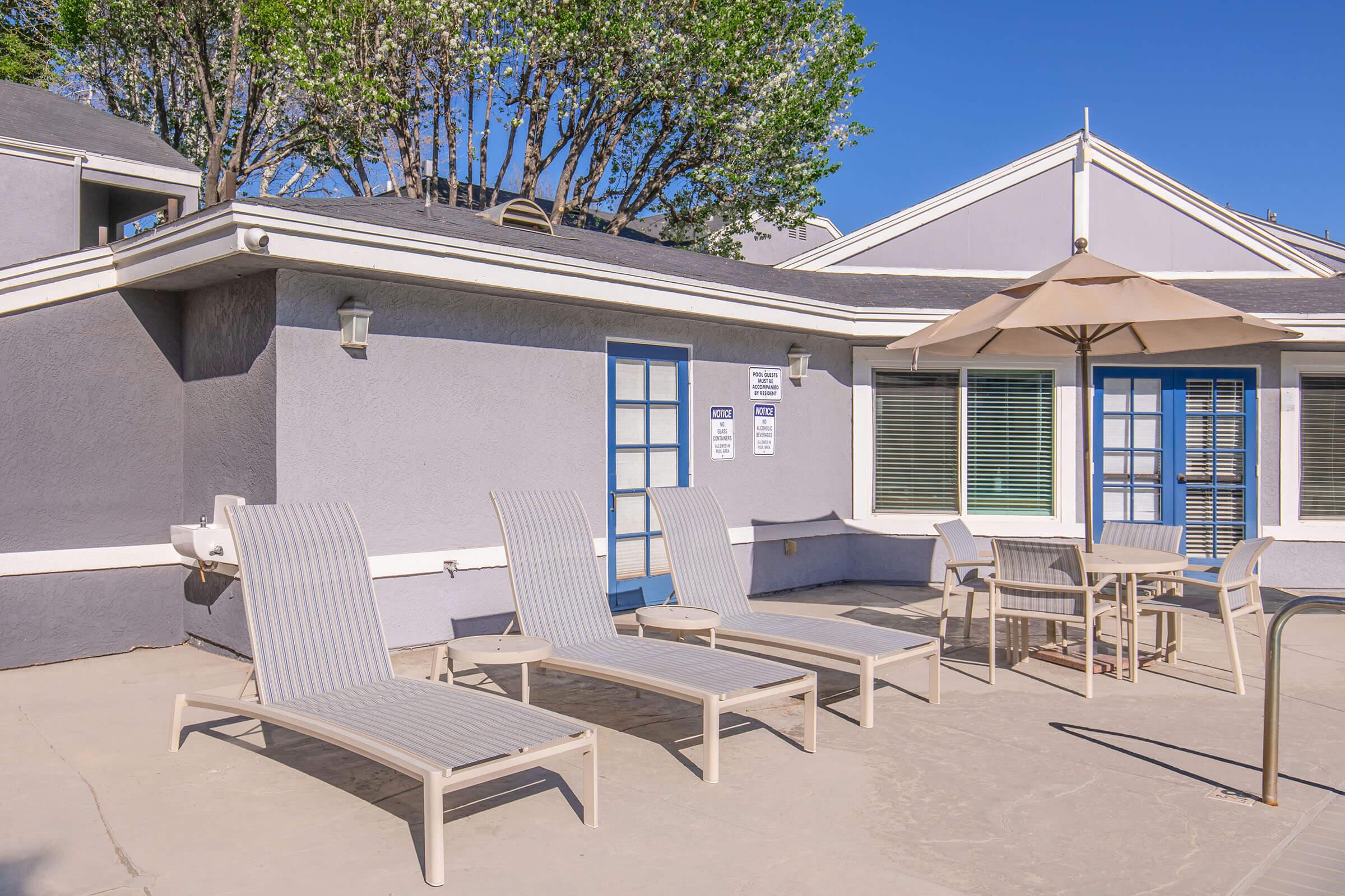 A sunny outdoor patio area featuring several lounge chairs, a table with chairs, and an umbrella. The walls are a light gray color, with blue doors and windows. Lush trees are visible in the background against a clear blue sky, creating a relaxing atmosphere.