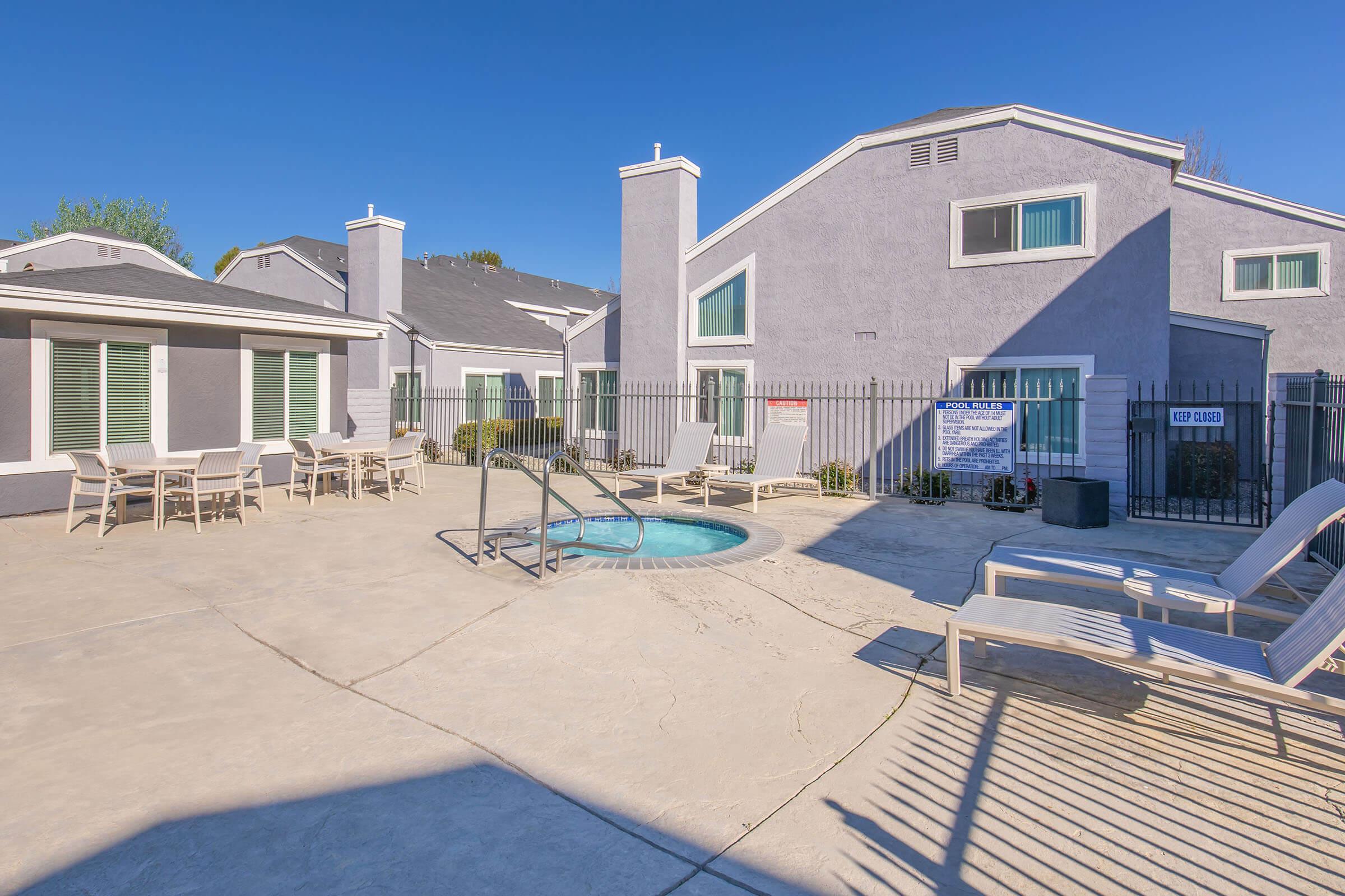 A sunny courtyard featuring a small circular hot tub surrounded by lounge chairs, with a fenced area and several buildings in the background. The buildings have modern architectural designs with large windows and pastel-colored exteriors.