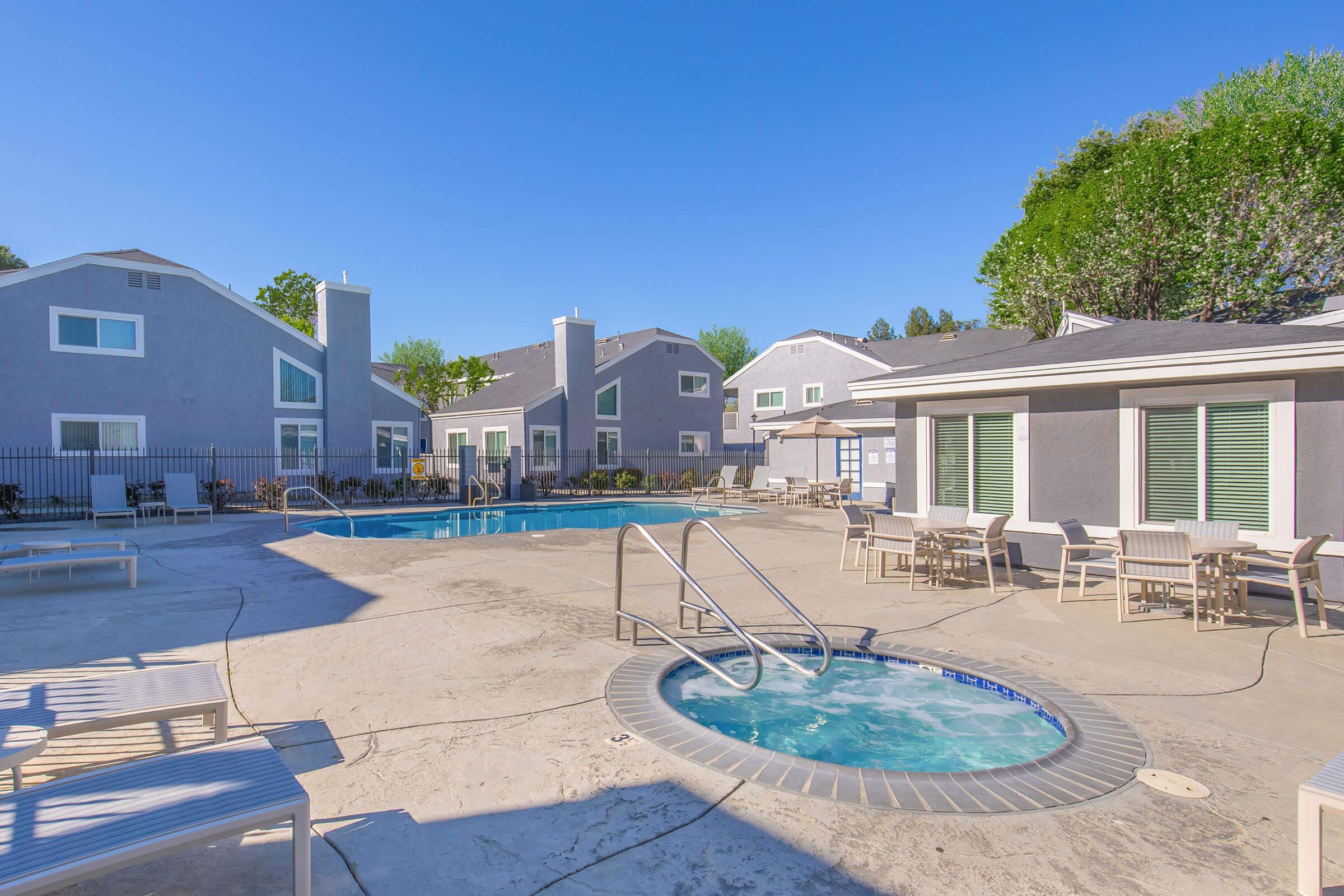 A sunny pool area featuring a large swimming pool and a hot tub. Surrounding the pool are lounge chairs and umbrellas. The backdrop includes several gray apartment buildings with green trees in the distance, creating a relaxing outdoor atmosphere.