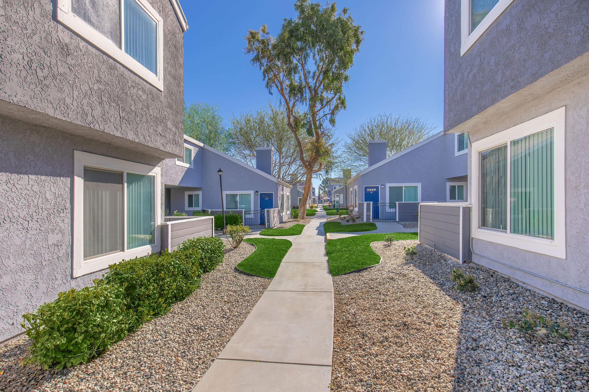 A pathway lined with grass and small shrubs leads through a residential area of modern buildings. The buildings have textured gray exteriors and large windows, surrounded by trees and clear blue skies. Sunlight bathes the scene, creating a bright and inviting atmosphere.