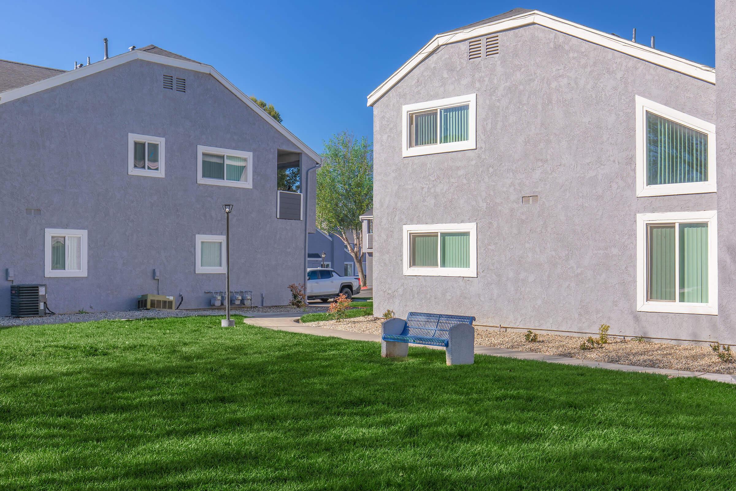 Two modern residential buildings with gray exteriors are situated across from each other. In the foreground, a blue bench sits on a well-maintained lawn with green grass. The scene is brightened by a clear blue sky, and there are small shrubs and landscaping elements around the area.