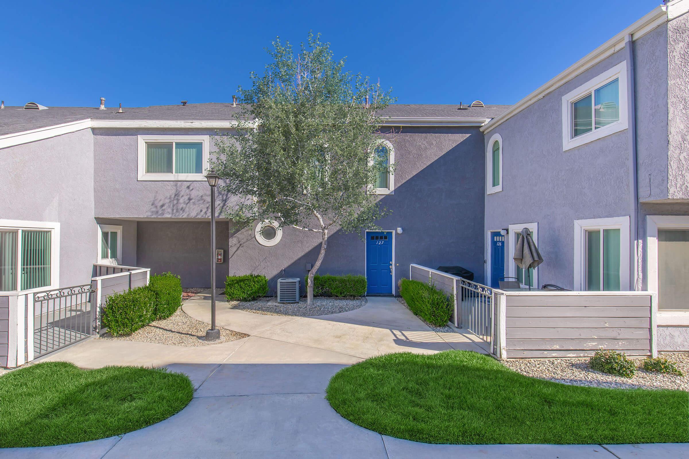 A landscaped pathway leads through a sunny courtyard with green grass, flanked by two modern apartment buildings. One building features a blue door and large windows, while the other has a neutral-colored facade and a small patio area. A tree provides shade in the center of the space.