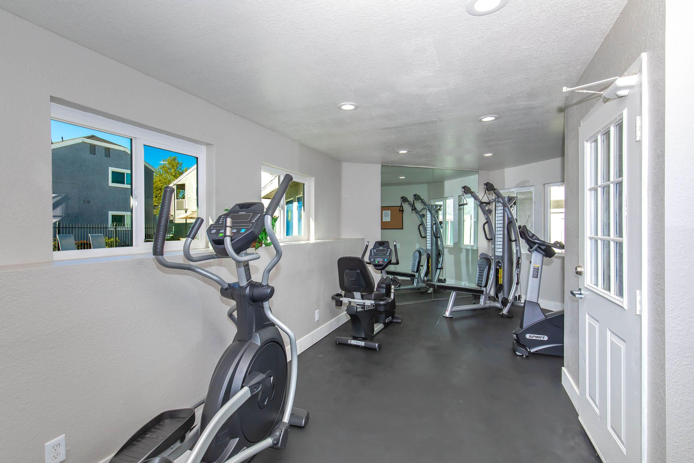 A modern gym space featuring various exercise equipment, including an elliptical machine, a stationary bike, and a weight machine. The room is well-lit with natural light from large windows, and has a clean, minimalist design. The entrance features a door leading outside.