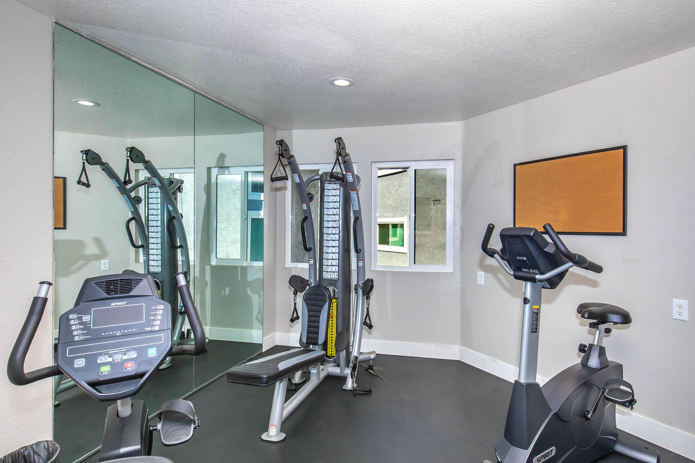 A small home gym featuring an exercise bike, a weight machine, and a mirror on one wall. The room has light-colored walls and a window providing natural light. A bulletin board is mounted on the wall, and the flooring is dark, creating a contrast with the lighter walls.