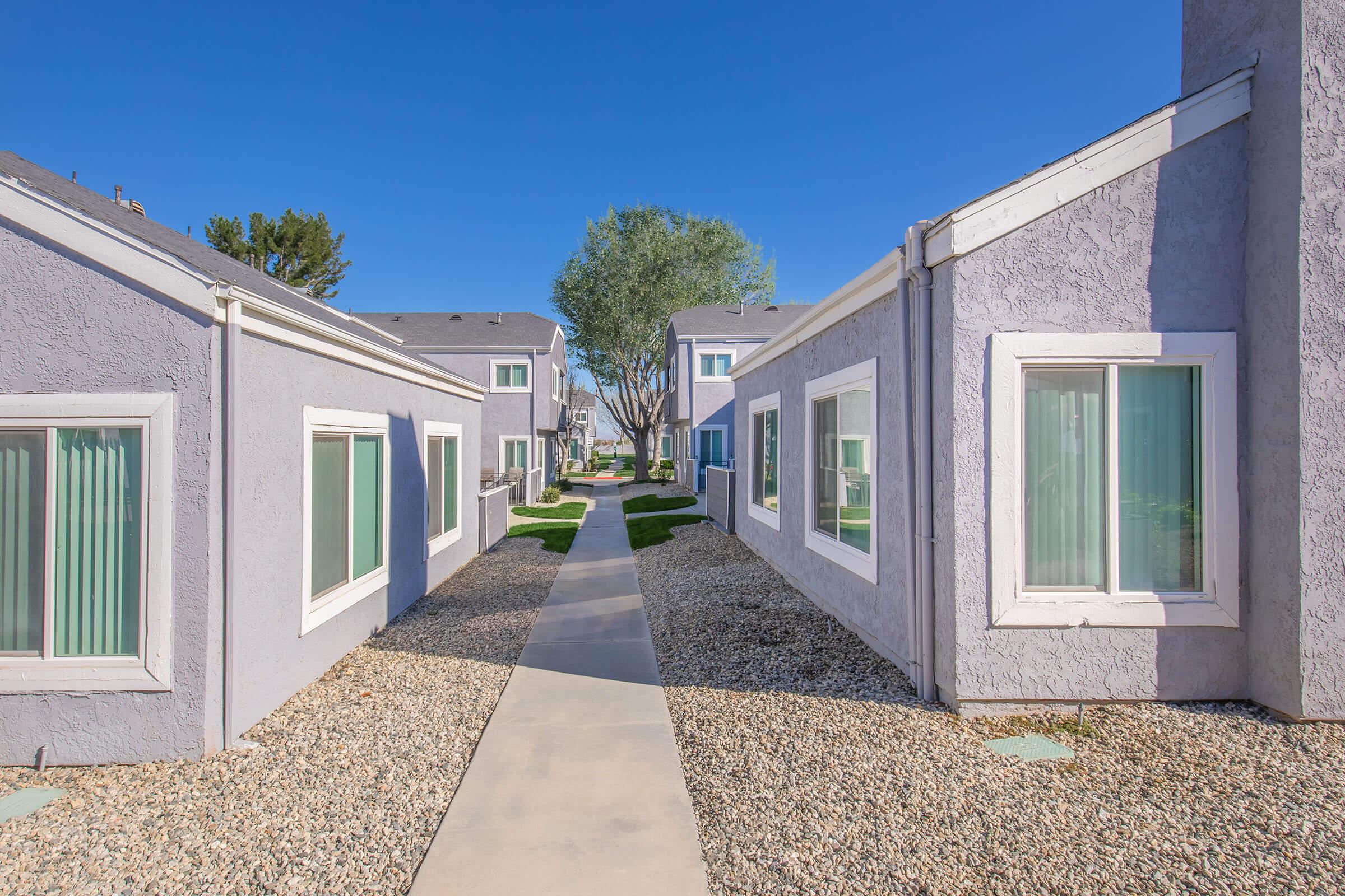 A clear pathway between two rows of residential buildings, featuring light purple exteriors and white trim. The walkway is lined with gravel, leading to greenery and trees in the background, under a bright blue sky.