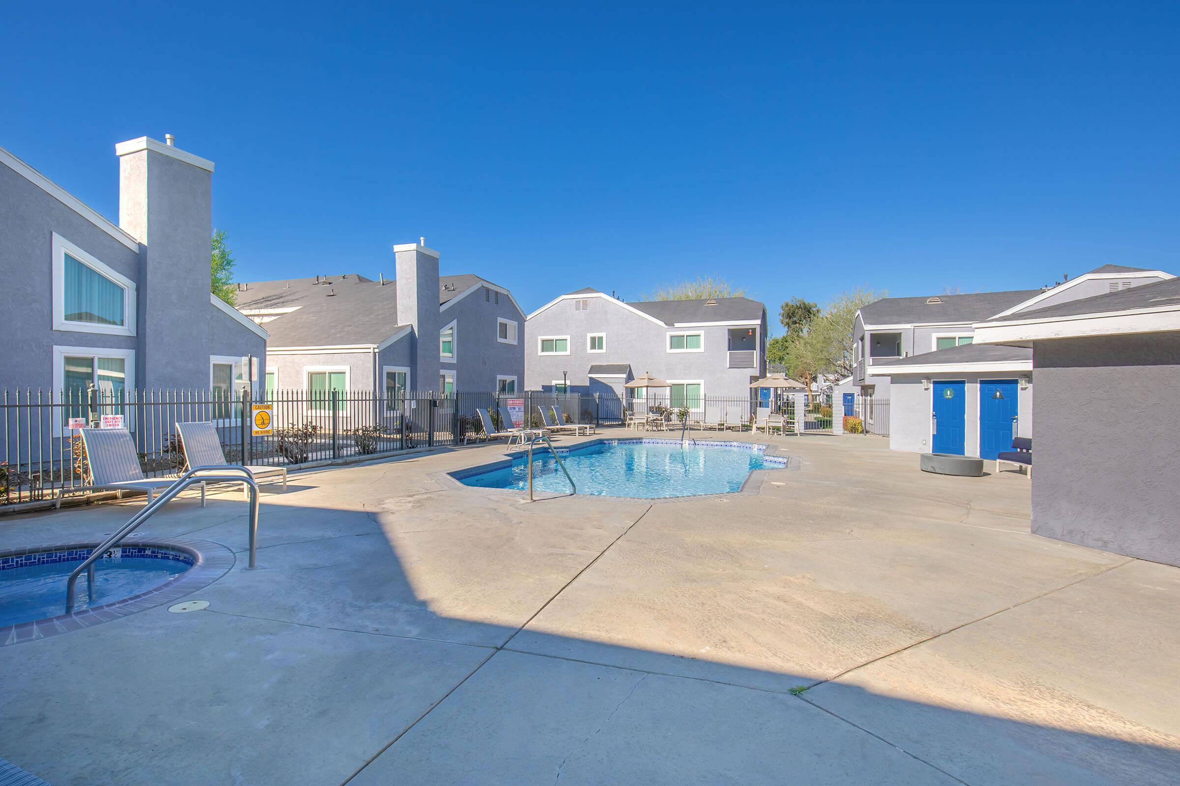 A sunny outdoor pool area featuring a rectangular swimming pool surrounded by lounge chairs. Gray buildings with white trim are visible in the background, along with a clear blue sky. The pool area is enclosed by a fence and includes a safety railing near the steps into the pool.