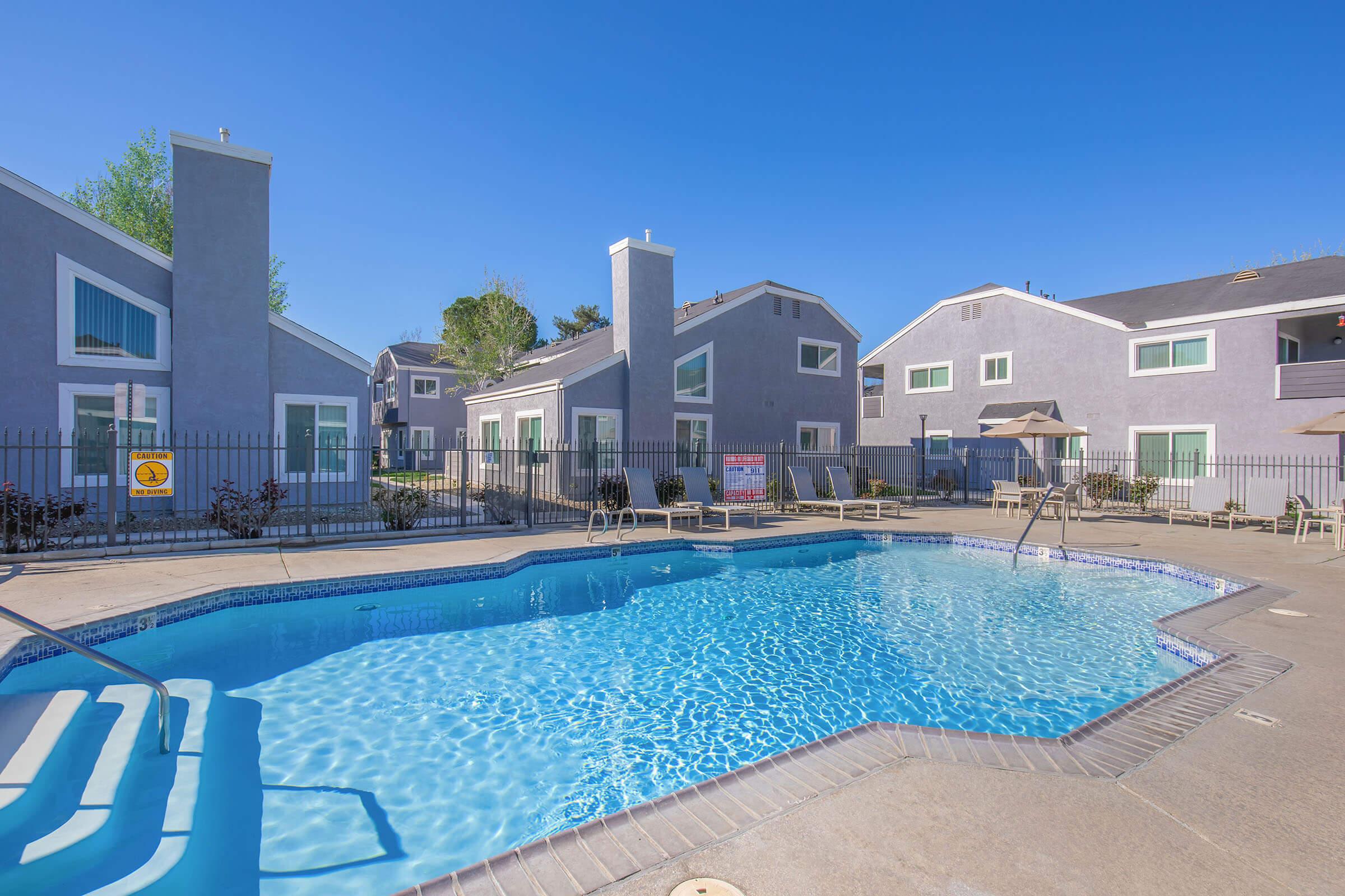 A clear blue swimming pool surrounded by a concrete patio. Lounge chairs are set up around the pool area. Modern, gray apartment buildings with green windows are in the background under a bright blue sky. A fenced area encloses the pool for privacy and safety.
