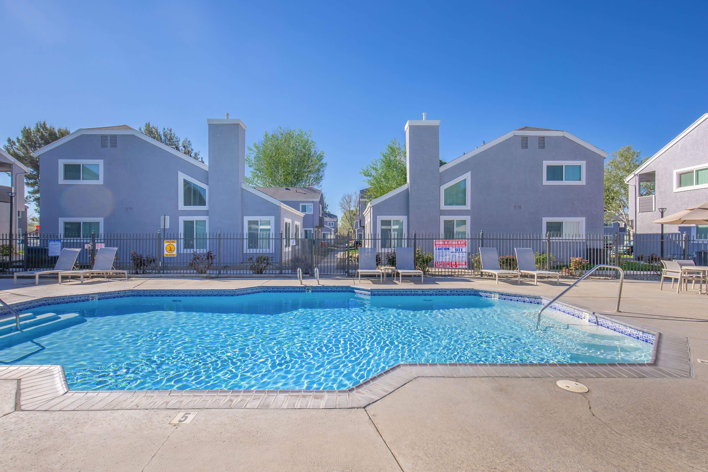 Clear blue swimming pool surrounded by lounge chairs, with gray apartment buildings in the background. Lush greenery adds to the inviting atmosphere under a bright blue sky.