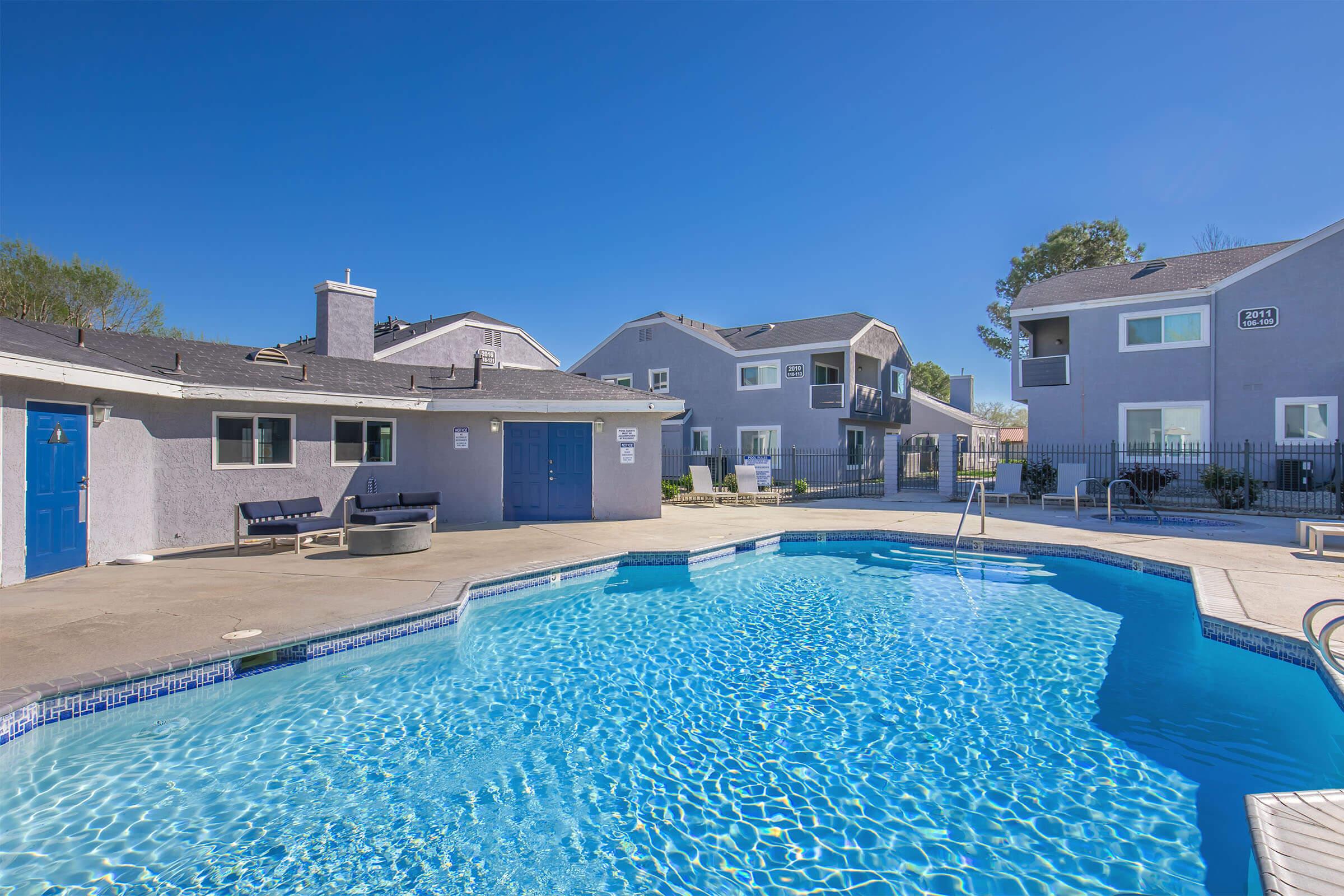 Swimming pool with clear blue water in a well-maintained area, surrounded by light-colored buildings. There are lounge chairs and a barbecue area nearby, under a bright blue sky. The setting is inviting and suitable for relaxation and leisure activities.