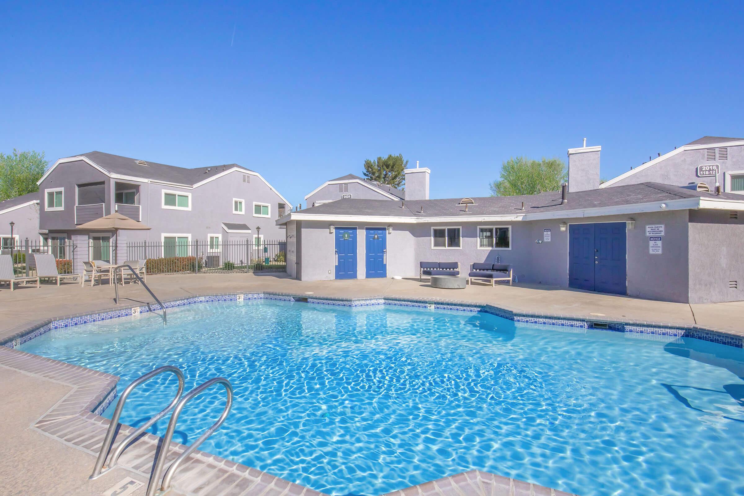 A clear blue swimming pool surrounded by lounge chairs and a concrete deck. In the background, there are several residential buildings with light-colored exteriors. The sky is bright and clear, creating a sunny atmosphere. A shaded area with tables is visible near the pool.
