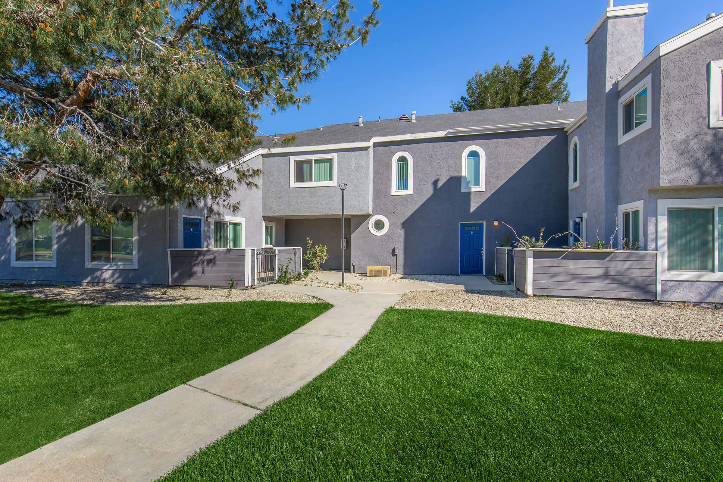 A modern building exterior featuring two residential units with blue doors, surrounded by lush green grass and a clear blue sky. A concrete pathway leads through the yard, bordered by landscaping. The structure has a contemporary design with gray walls and several windows.