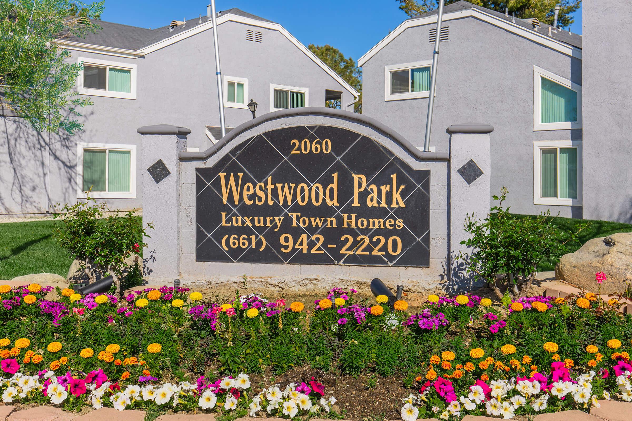 Sign for Westwood Park Luxury Town Homes, located at 2060, featuring a dark stone design with gold lettering. The sign displays a phone number and is surrounded by colorful flower beds with vibrant blooms, set against a backdrop of modern multi-unit buildings and greenery.