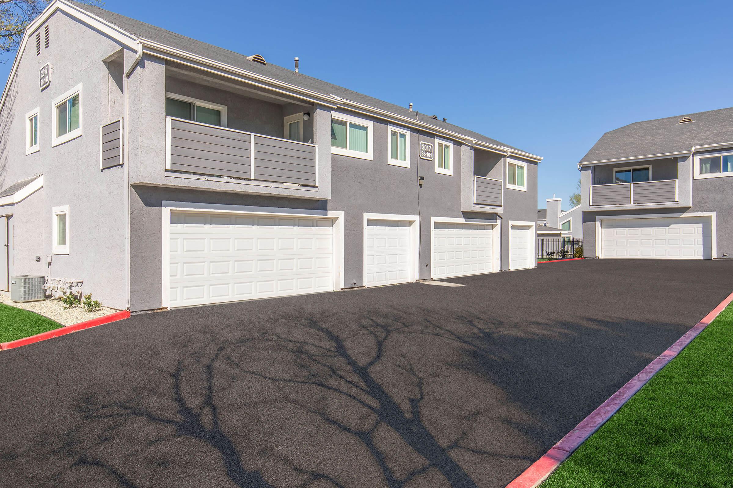 A residential building with multiple units, featuring grey exterior walls and garages with white doors. The area has a newly paved driveway, and the shadow of a nearby tree is visible on the pavement. Clear blue sky in the background.