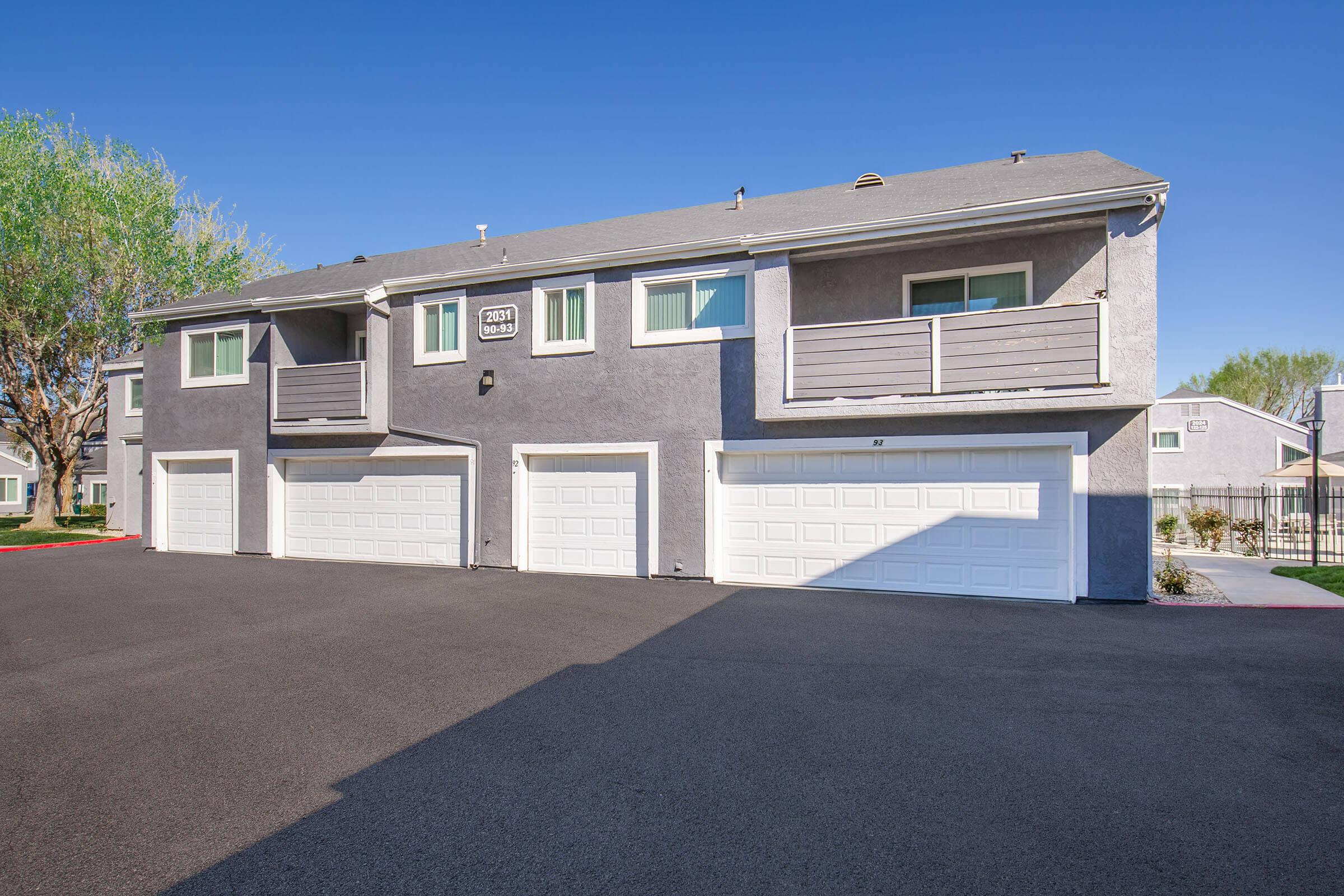 A multi-unit residential building with multiple garages below. The building features light gray exteriors, windows with white frames, and a clear blue sky overhead. The driveway is paved and well-maintained, with green trees visible in the background.