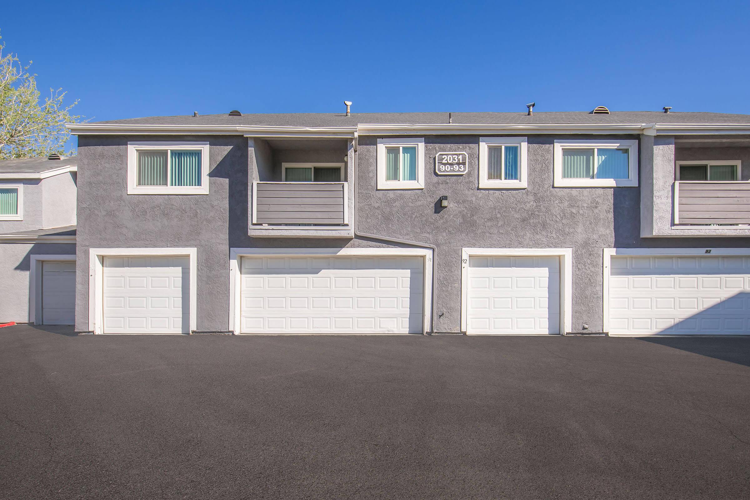 A row of residential townhouses featuring multiple garage doors at ground level. The exterior is painted in shades of gray, with large windows and balconies visible on the upper floors. The sky is clear and blue, providing a bright backdrop for the building. The area in front of the garages is paved.