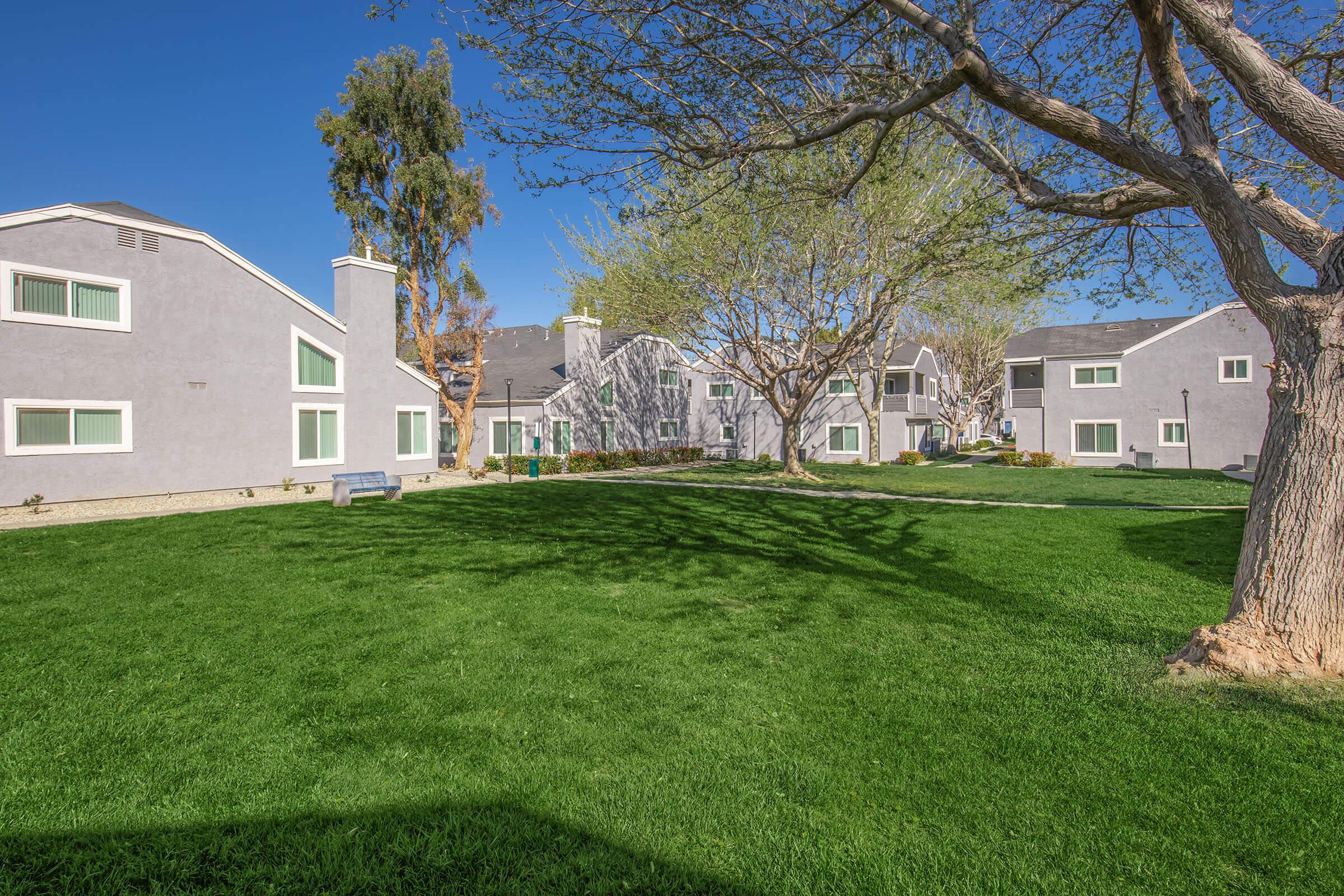 A sunny residential area featuring gray multi-unit buildings surrounded by well-maintained green lawns and trees. The landscape includes a spacious outdoor area with a clear blue sky, creating a peaceful and inviting atmosphere.