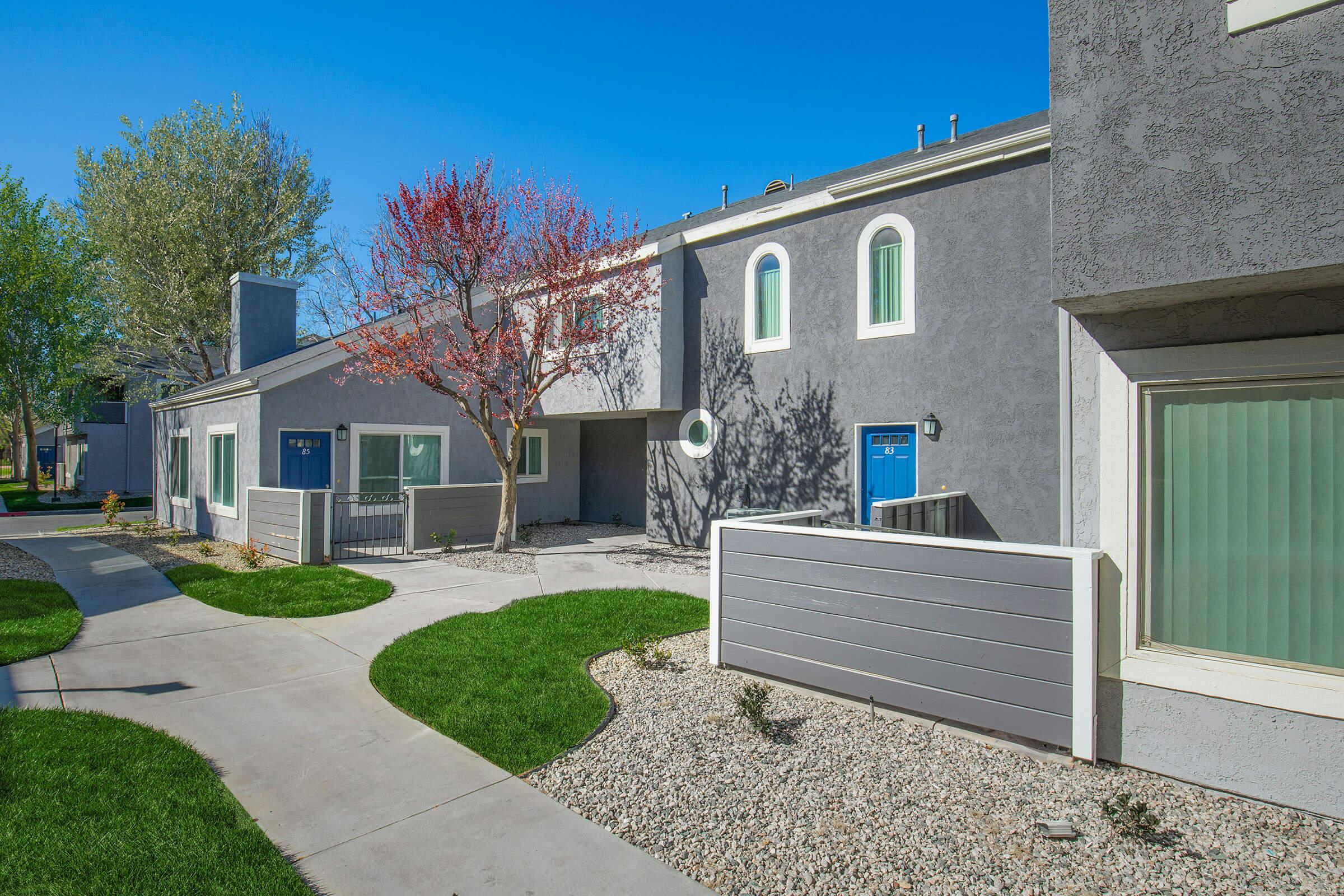 A pathway leading through a well-maintained residential area featuring modern gray buildings with blue doors. A flowering tree adds color, and manicured lawns with rock landscaping complement the scene under a clear blue sky.