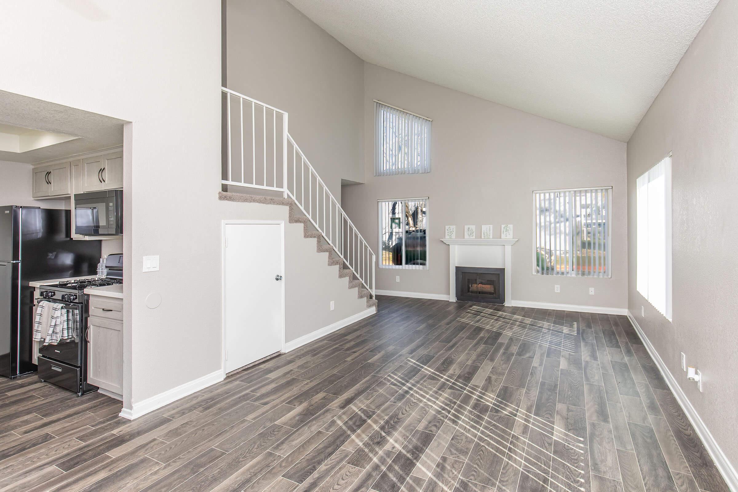 Spacious living area with high ceilings, featuring large windows that allow natural light, a stairway leading to an upper level, and a modern fireplace. The adjacent kitchen area is visible with dark cabinetry and appliances, creating a welcoming and airy atmosphere.