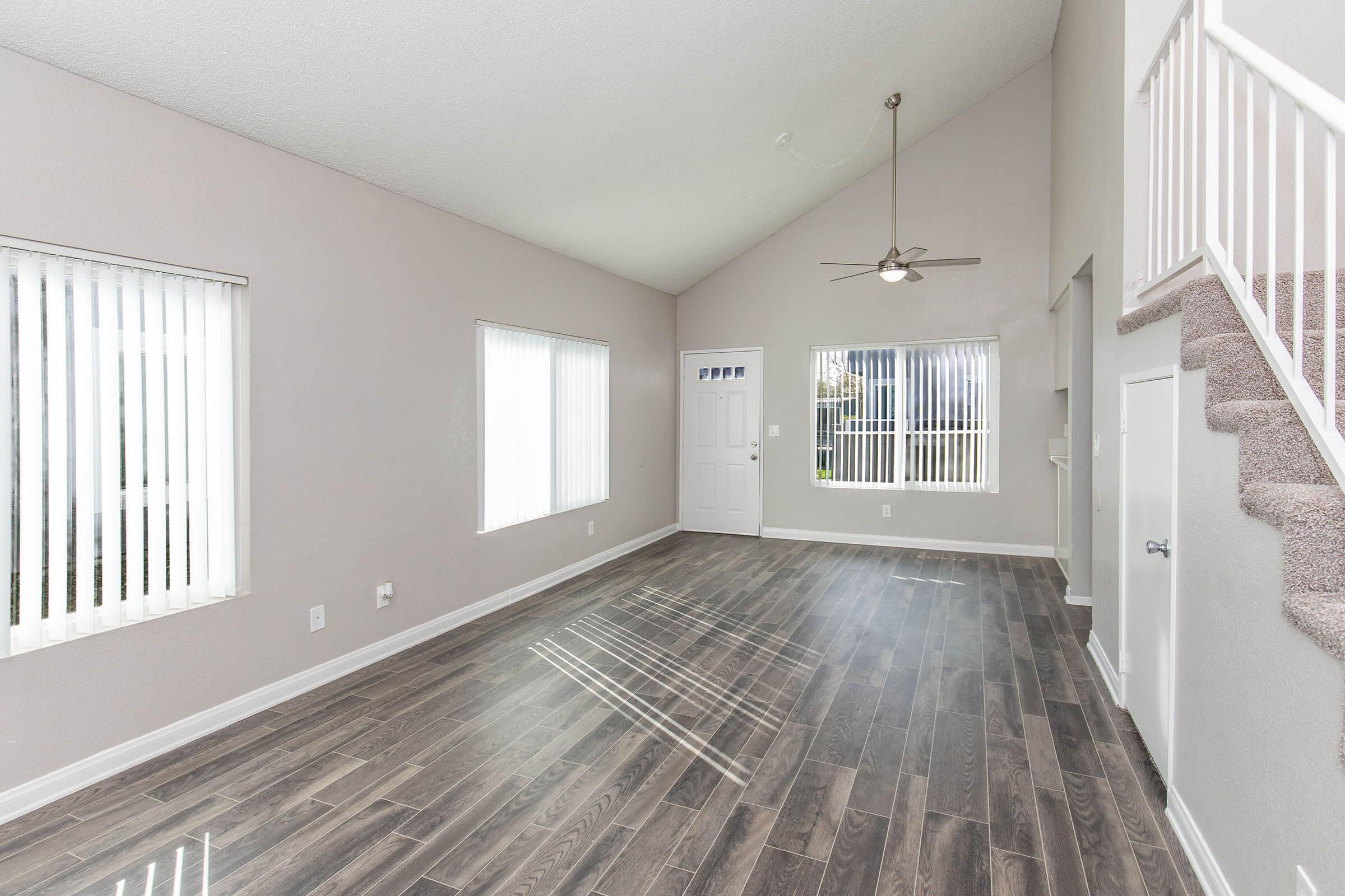 Spacious, well-lit living room with high ceilings, featuring large windows with vertical blinds, hardwood-style flooring, and a ceiling fan. A staircase is visible on the right, leading to an upper level. The room has neutral-colored walls and a front door visible.