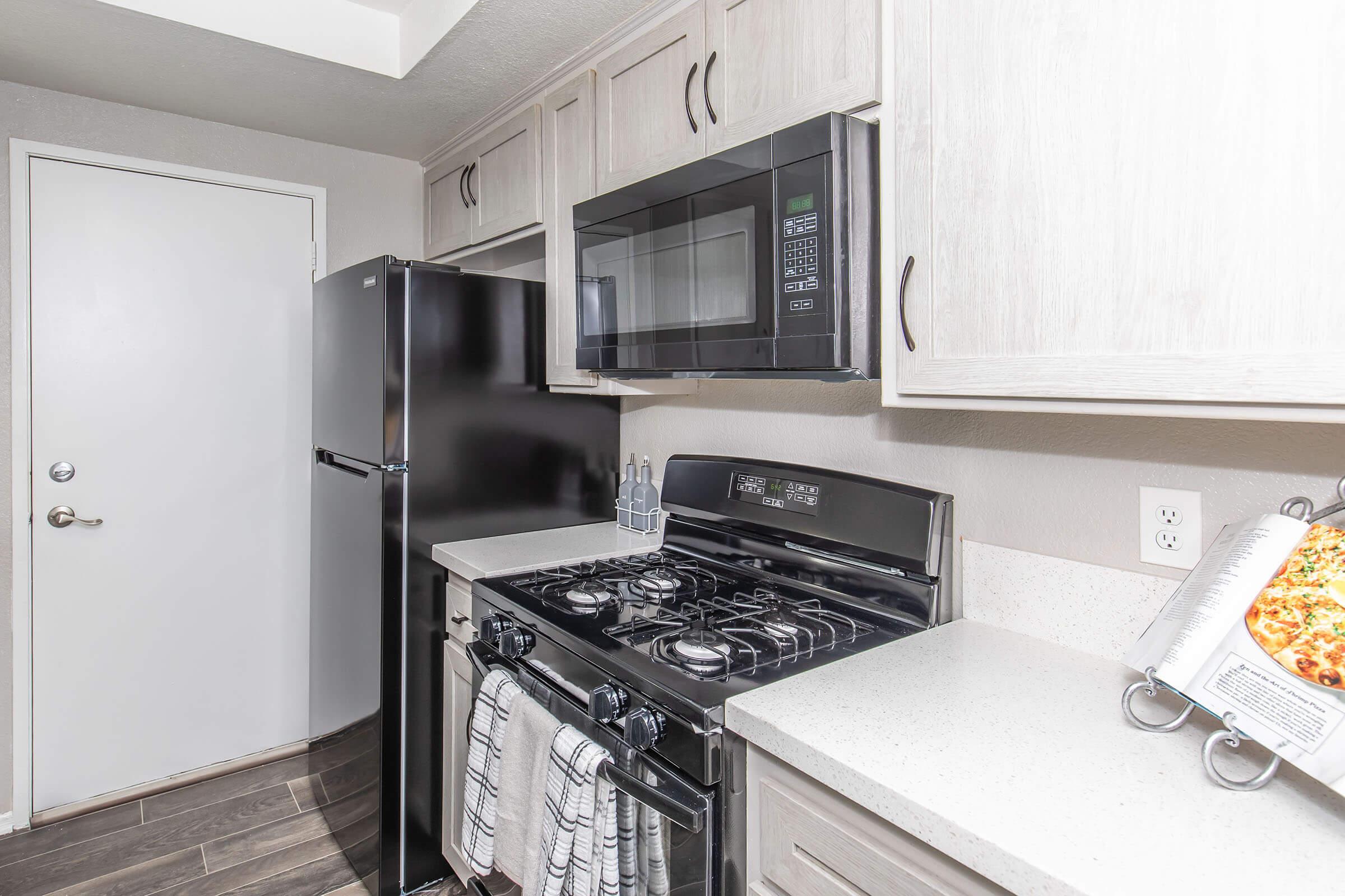 A modern kitchen featuring light wood cabinetry, a black refrigerator, an over-the-range microwave, and a gas stove. The countertop is a light-colored material, and a dish towel hangs on the stove. A door leads out of the kitchen, and a cookbook with a stand sits on the counter, showcasing a dish.
