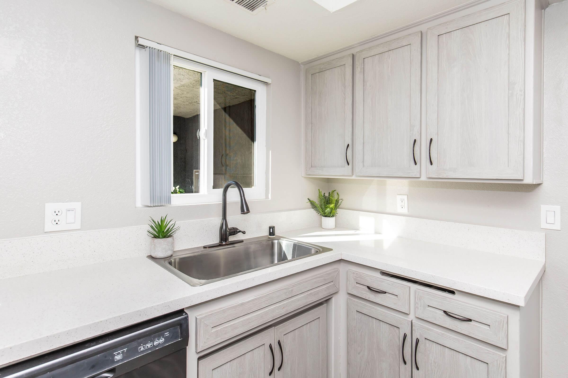 Modern kitchen featuring light gray cabinetry, a stainless steel sink, and a countertop with a clean, minimal design. Natural light enters through a window, and small potted plants are placed on the countertop for a touch of greenery. The overall aesthetic is sleek and contemporary.