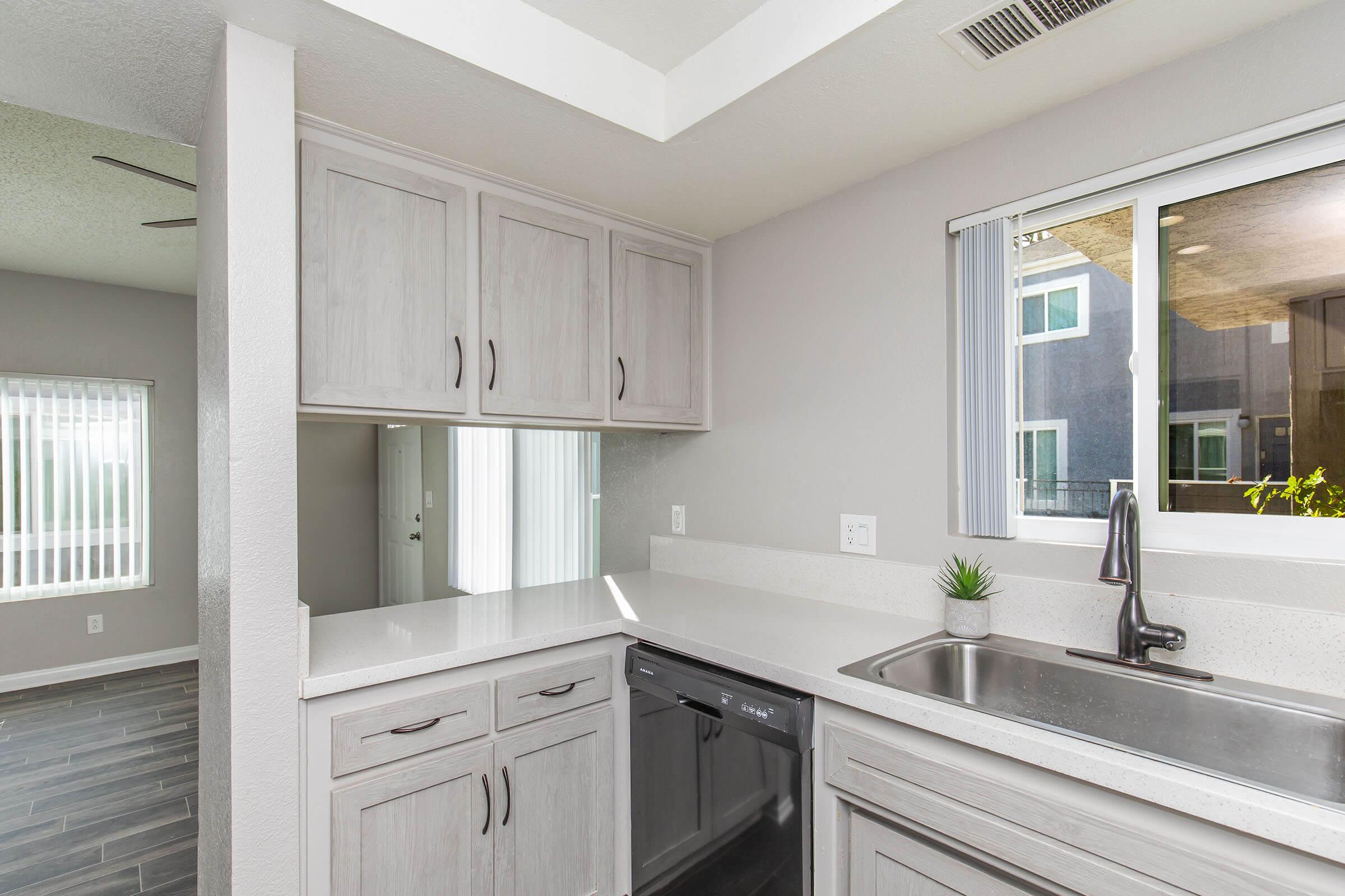 Modern kitchen with light gray cabinets and a white countertop. Stainless steel sink with a sleek faucet, dishwasher underneath the counter, and a small plant for decoration. A window allows natural light to enter, showcasing an open layout that connects to a living area with large windows.