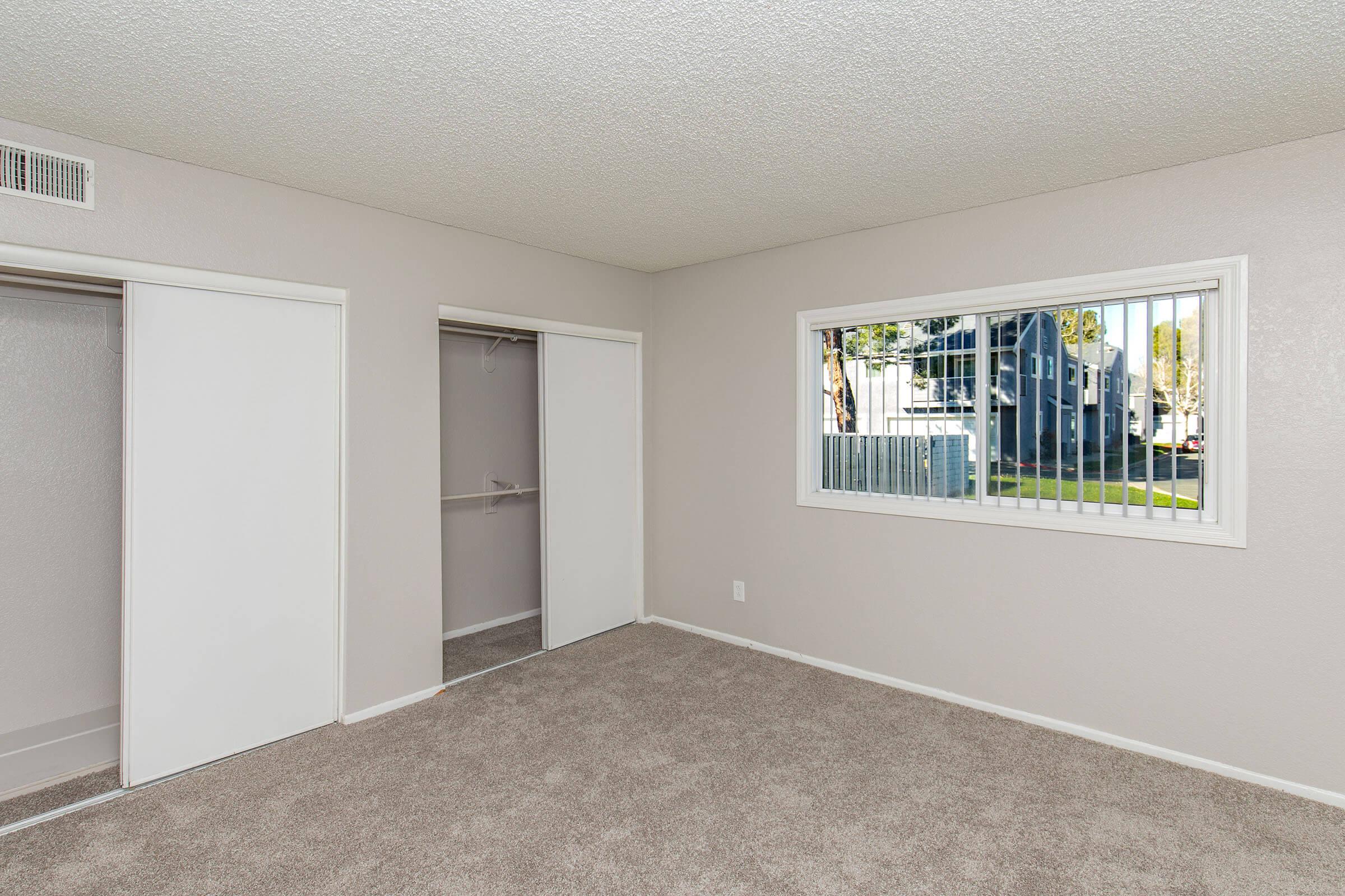 A light-colored room with beige carpet, featuring a large window with vertical blinds and two closet doors. The walls are painted a soft gray, and the room is well-lit, showing a view of the outside. The overall space appears clean and empty, ready for furnishing.