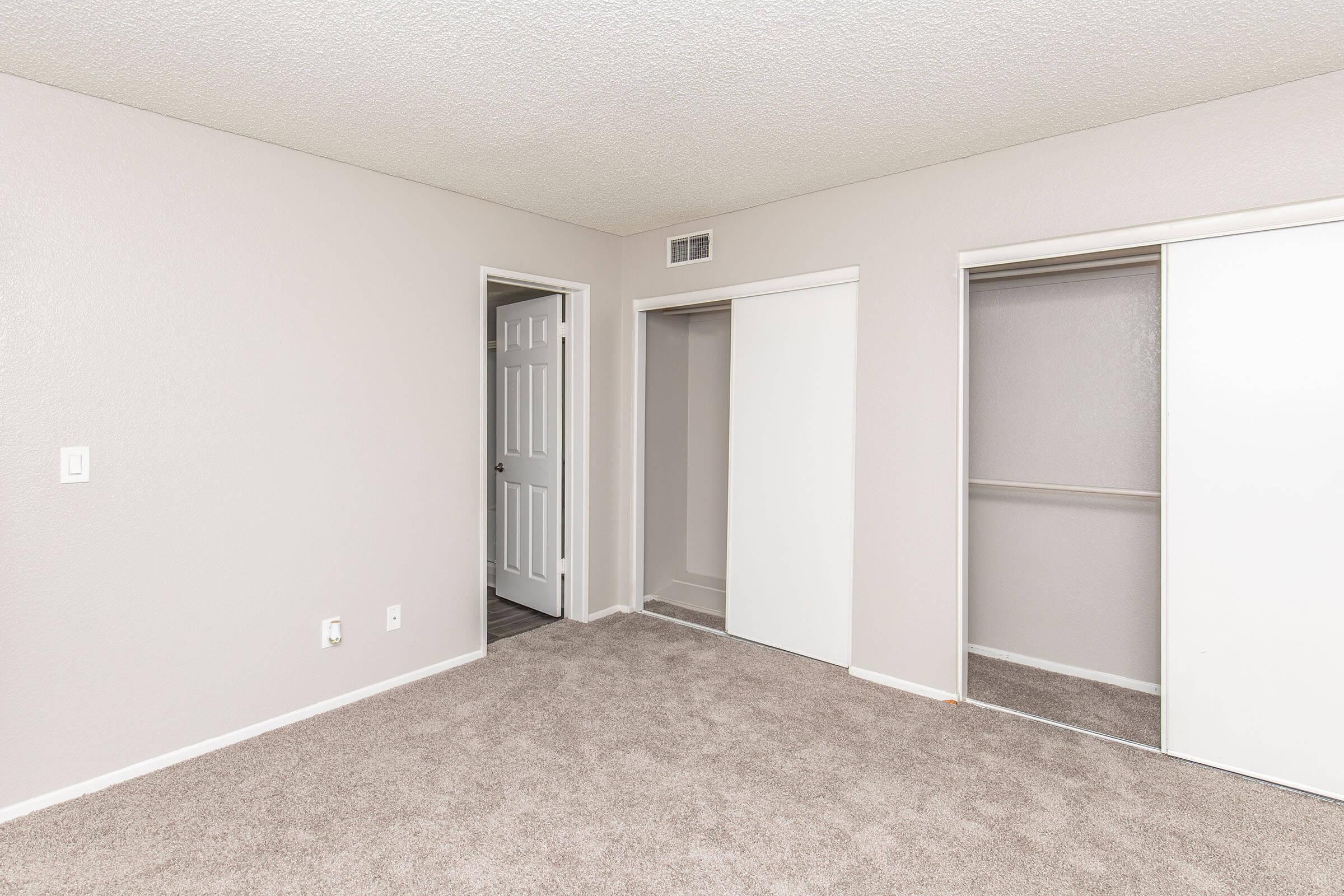 A spacious, well-lit room with light beige carpet and neutral-colored walls. There is a doorway leading to another room and a closet with sliding doors on the right. The room appears clean and empty, ideal for various decorative styles.
