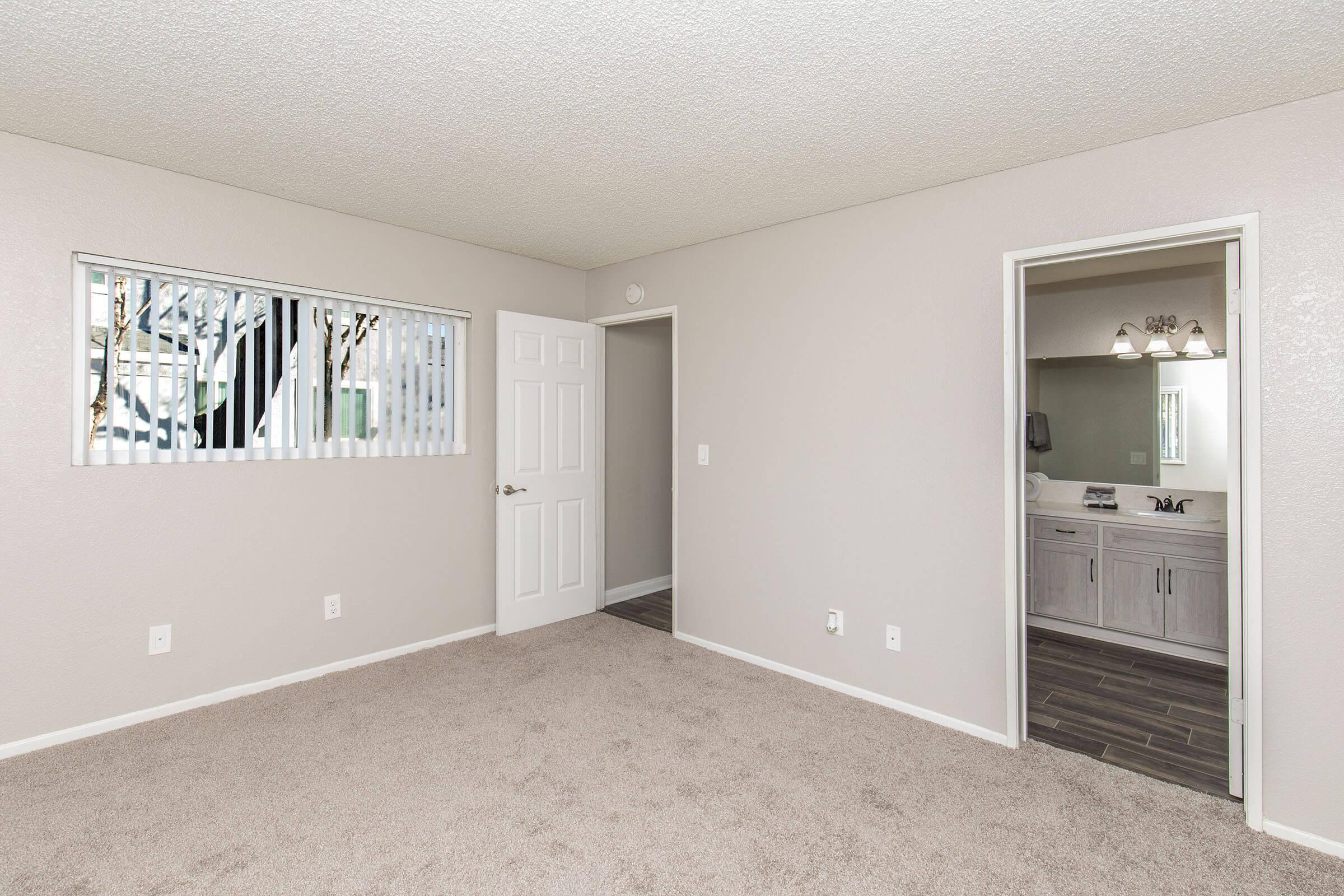 A bright, empty room with beige carpet and light gray walls. A window with vertical blinds is on the left, and a doorway leads to a bathroom on the right. The bathroom features a mirror and modern fixtures. The overall space is clean and uncluttered, suitable for various design styles.