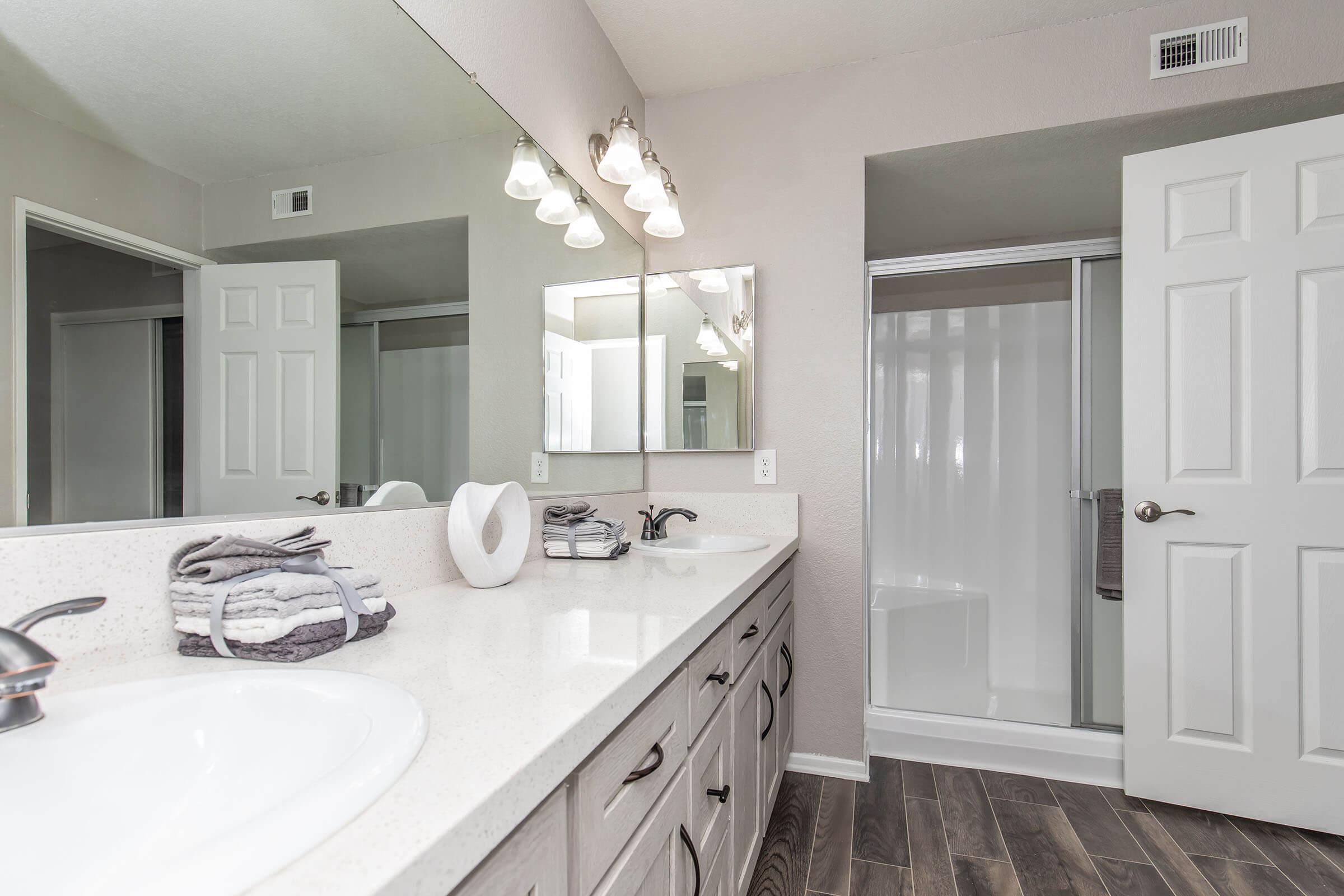 A modern bathroom featuring a double vanity with a spacious countertop, neatly arranged towels, and a large mirror. The room has a shower area behind a glass door and light fixtures above the mirror. The walls are painted in neutral tones, and the floor has a wood-like finish.