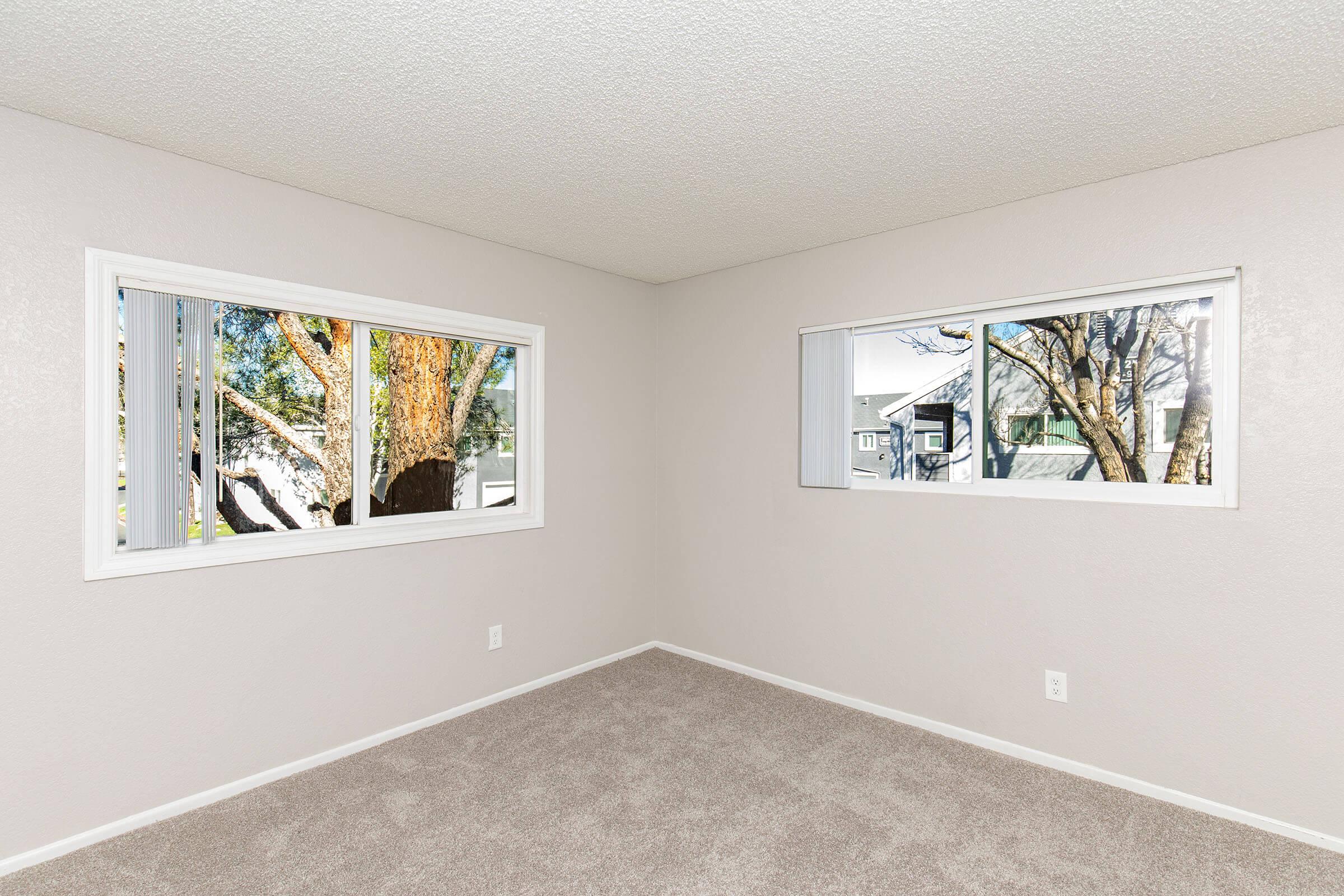 Empty room with light gray walls and plush carpet. Two large windows provide natural light, showcasing trees outside. The space appears clean and freshly painted, suitable for various uses.