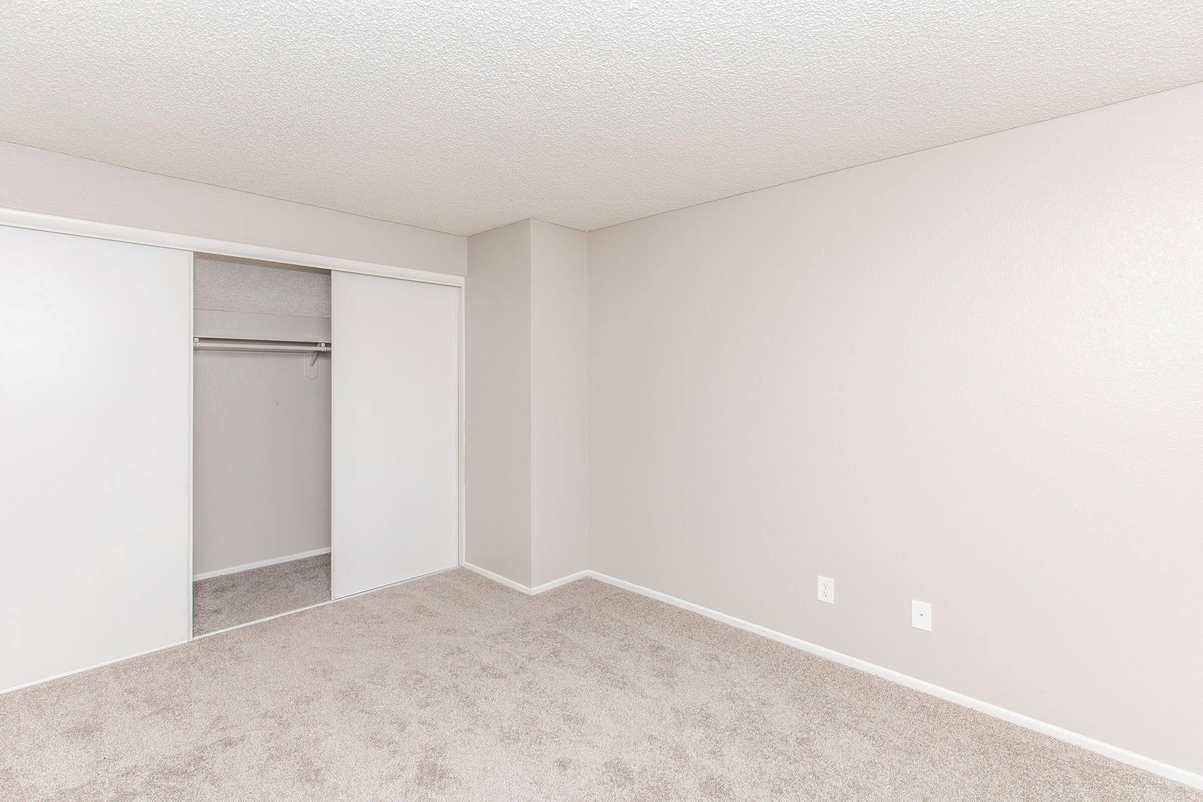 A spacious, empty room with light beige carpet and beige walls. A closet with sliding white doors is visible on the left side, and the room has no furniture. The corner of the room features a plain wall, contributing to a clean and minimalistic aesthetic.