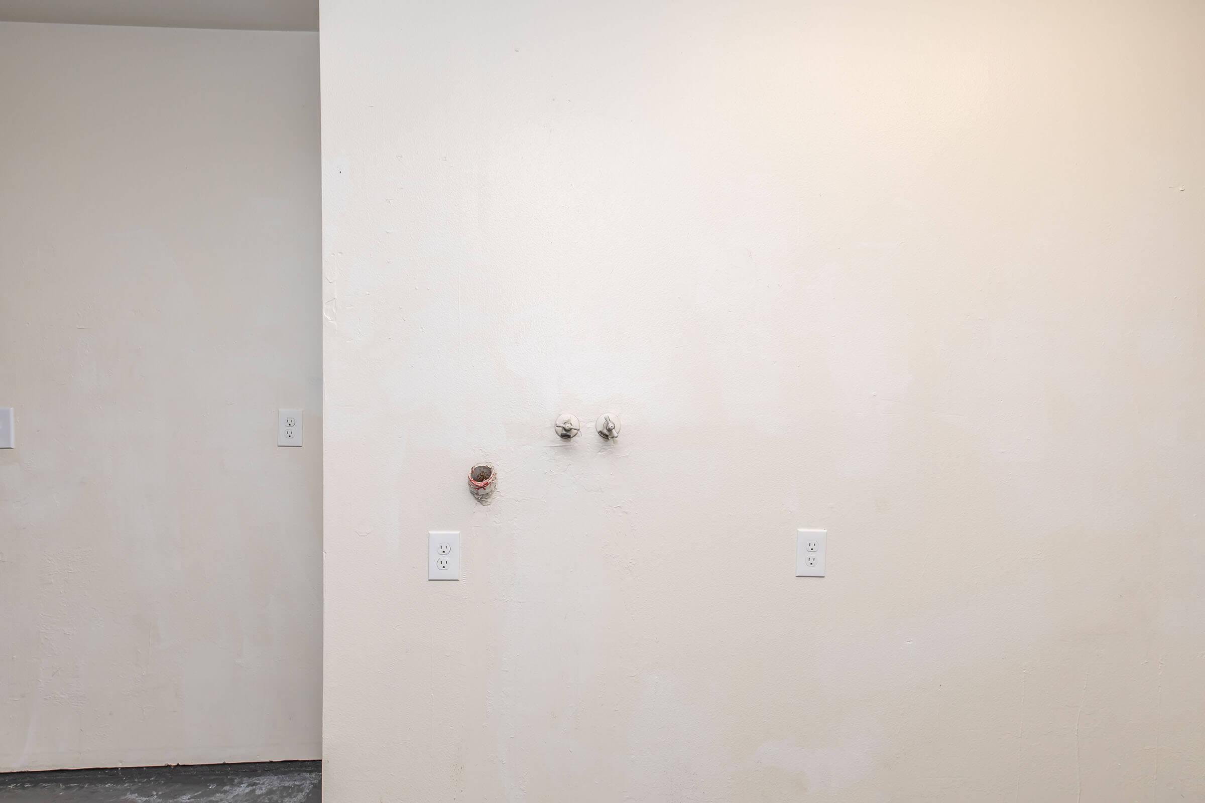 A plain, unfinished wall with two visible plumbing fixtures and an electrical outlet. The wall is light-colored and smooth, with minimal texture, suggesting it may be in a residential or commercial space under renovation. An adjacent wall can be seen to the left, featuring additional outlets.
