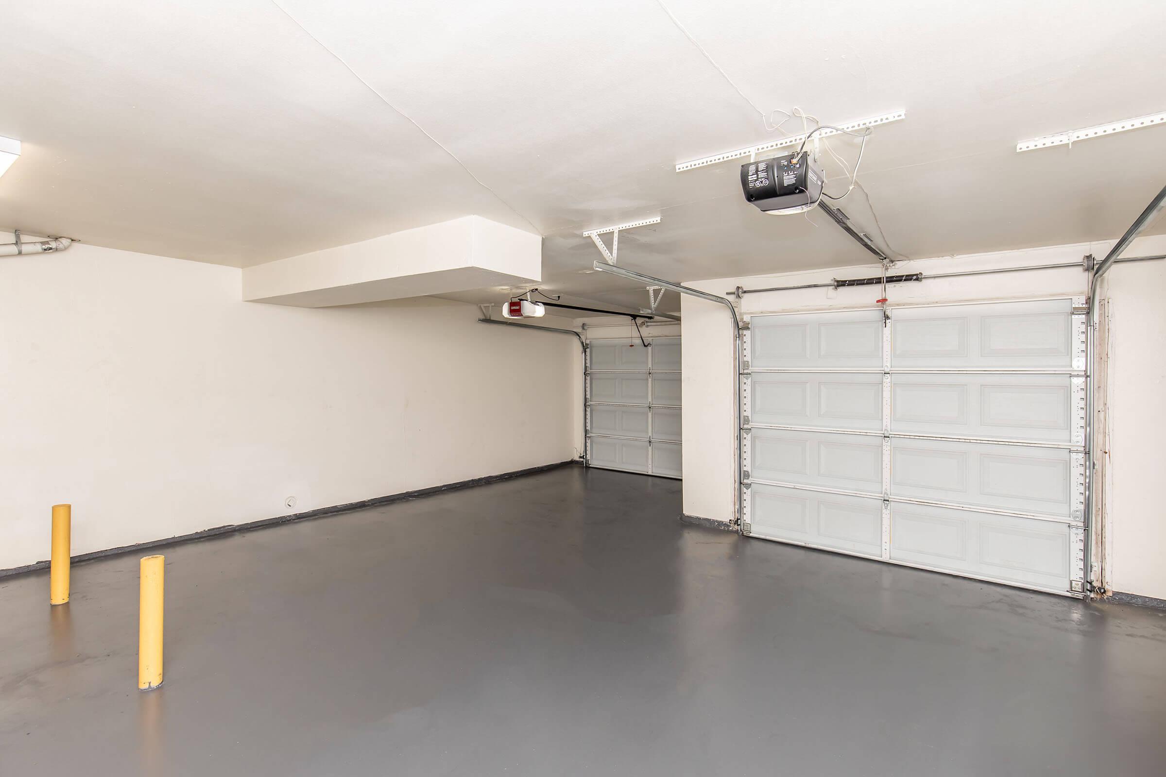 Spacious, empty garage with a smooth gray floor and two overhead garage doors on the left. There are yellow safety poles near the walls, and ceiling lights provide bright illumination. The walls are painted a neutral color, contributing to a clean and uncluttered appearance.