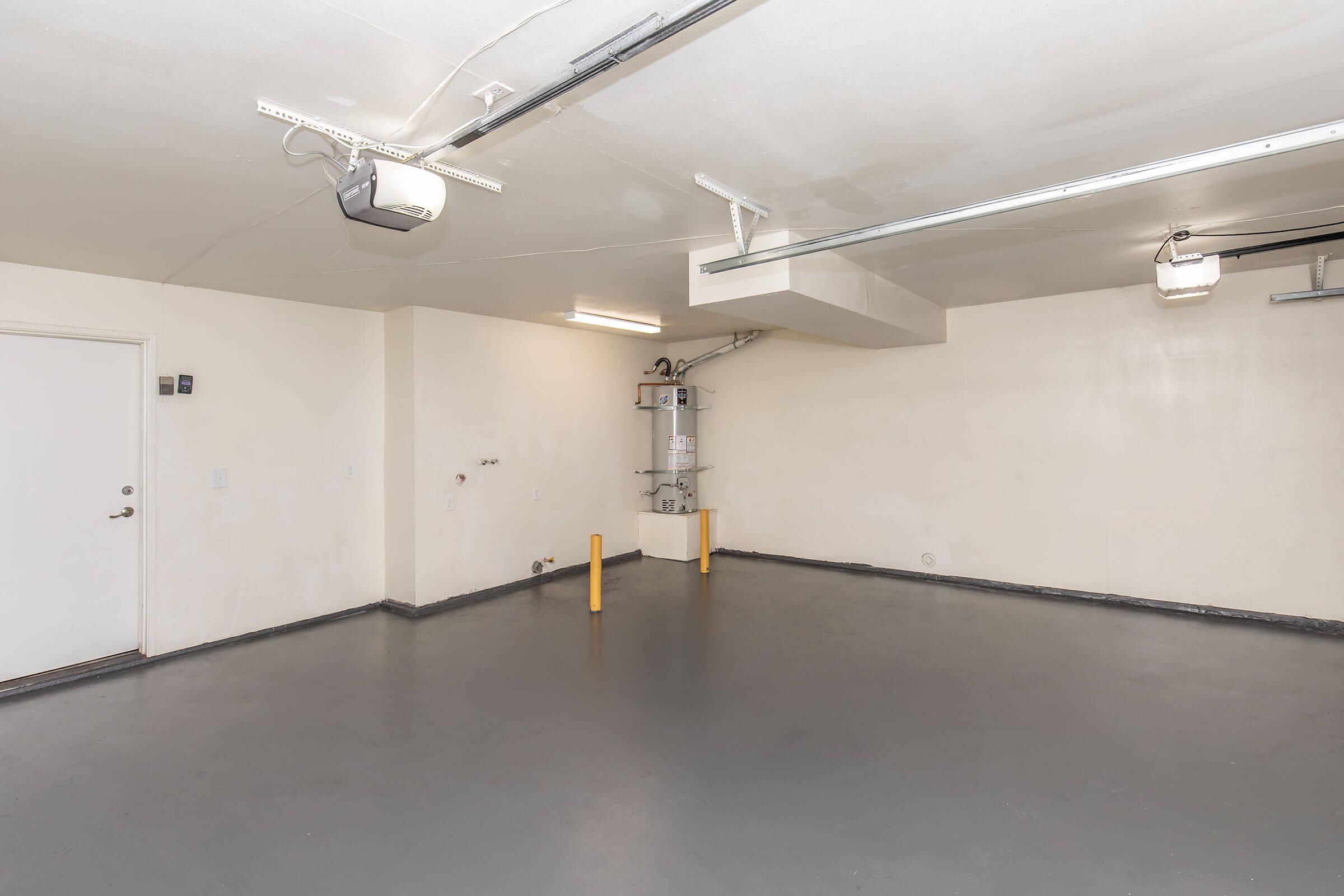 An empty, clean garage space with light gray walls and a smooth concrete floor. A single door is visible on one wall, and there are two ceiling-mounted garage door openers. The area is bright and uncluttered, featuring minimal furnishings and a neutral aesthetic.