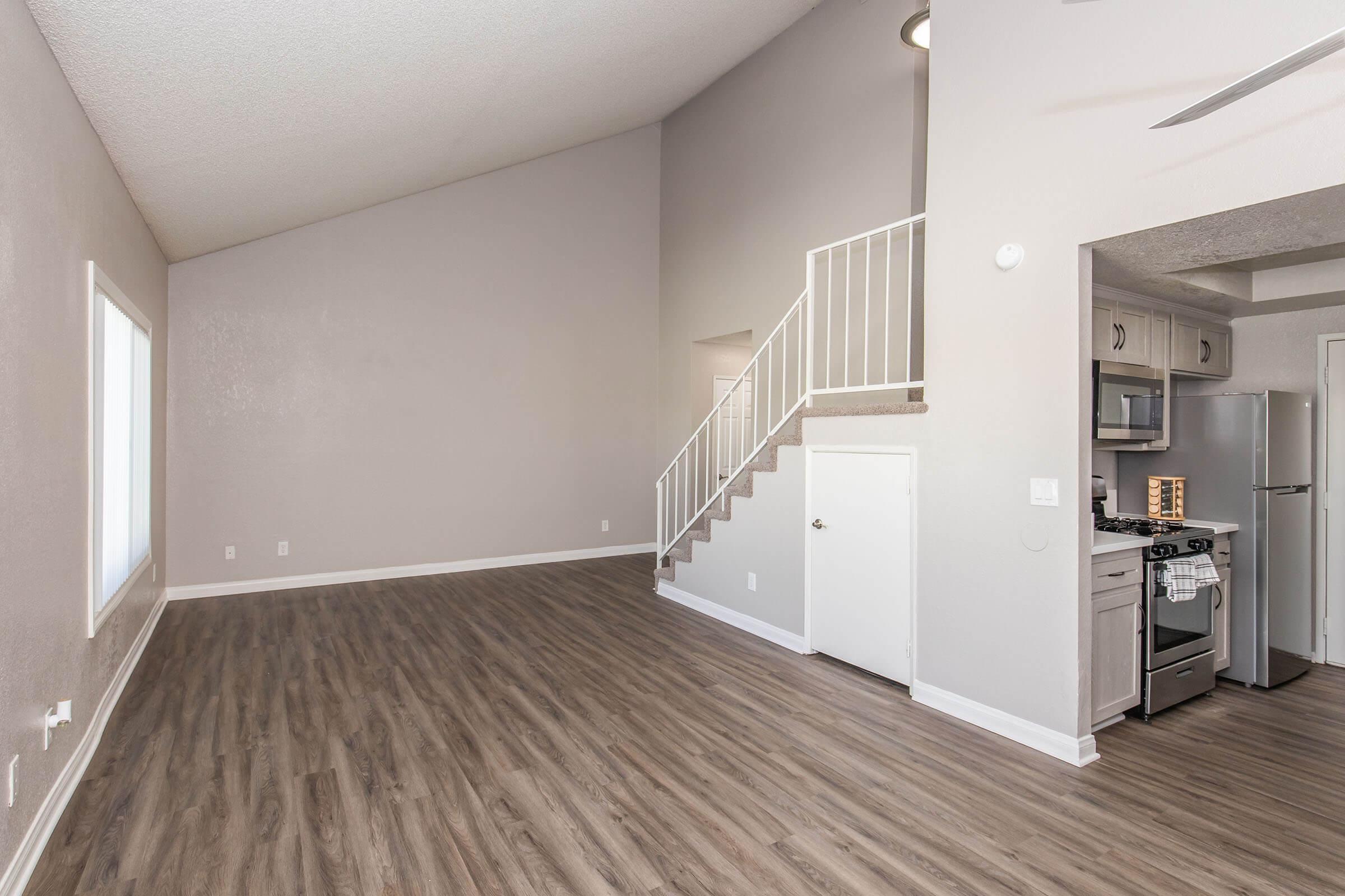 Interior view of a modern, spacious living area with high ceilings. The room features light-colored walls, hardwood flooring, and a staircase leading to an upper level. A kitchen area is visible, equipped with stainless steel appliances, and a small door leads to a storage space or closet. Natural light comes through a large window.