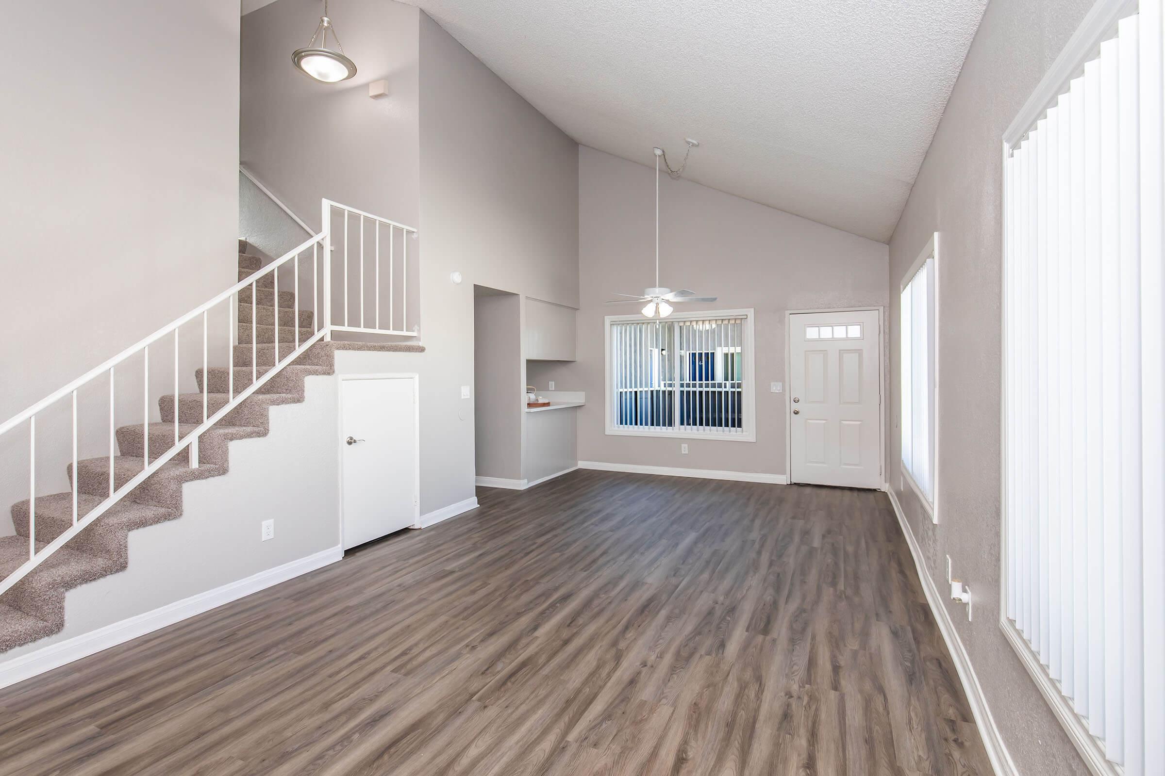 Spacious living area featuring high ceilings and large windows, with a staircase leading to the upper floor. Light-colored walls and modern wood-like flooring create a bright atmosphere. The open kitchen area is visible in the background, complemented by a door leading outside.
