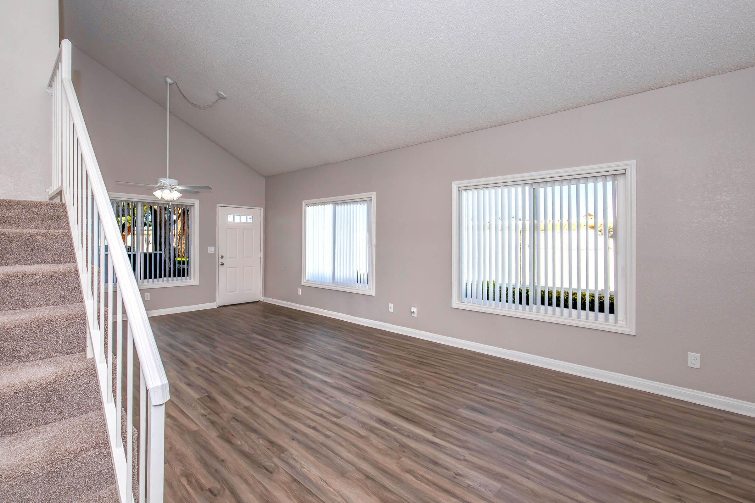 A spacious living room featuring light-colored walls and large windows with vertical blinds. There is a staircase to the left and vinyl flooring throughout. A ceiling fan hangs from the ceiling, adding to the airy atmosphere. The front door is visible, enhancing the room's natural light.