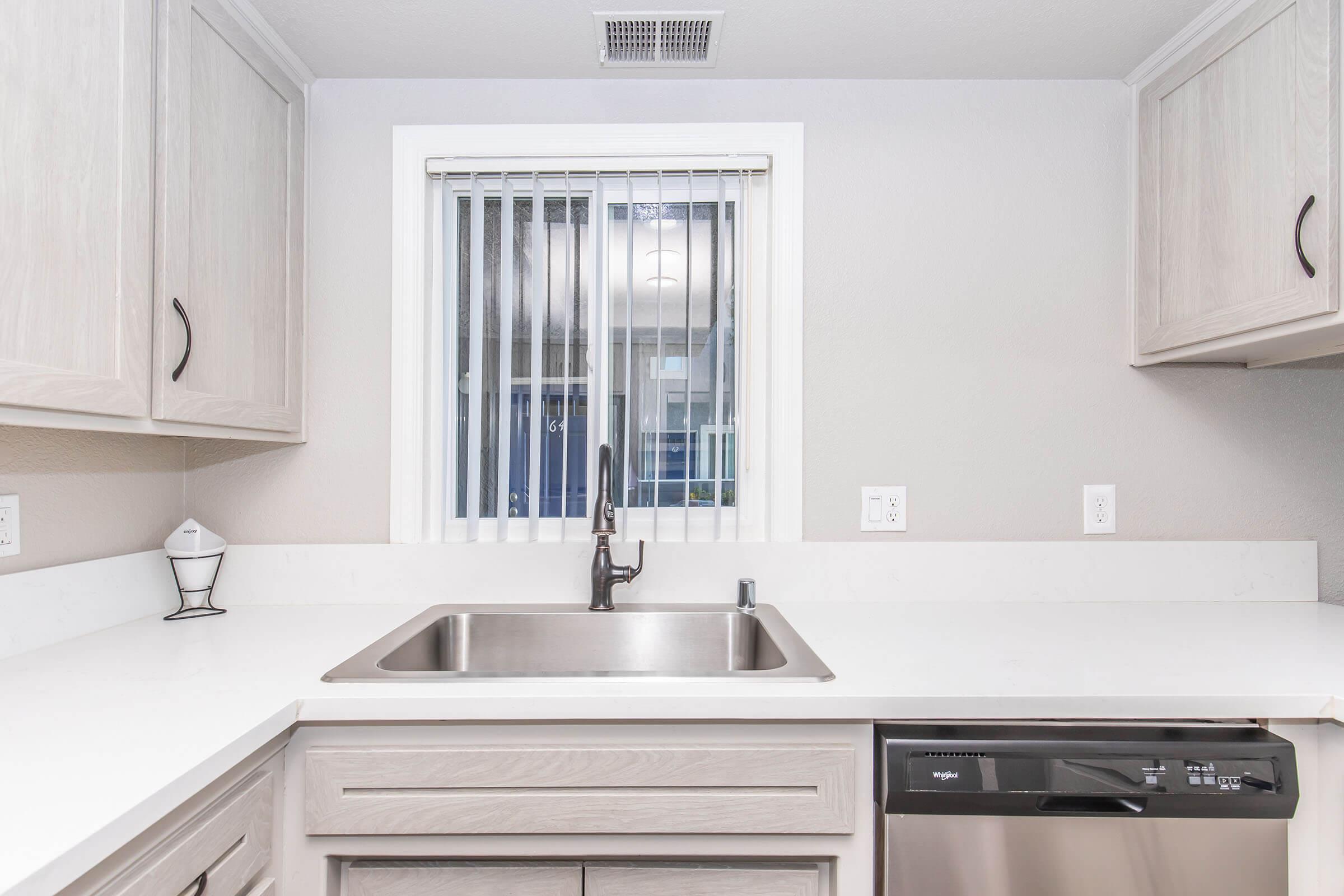 A modern kitchen featuring a stainless steel sink and dishwasher, light-colored cabinetry, and a window with blinds. The countertop is smooth and white, creating a bright and clean space. The overall aesthetic is contemporary and functional.