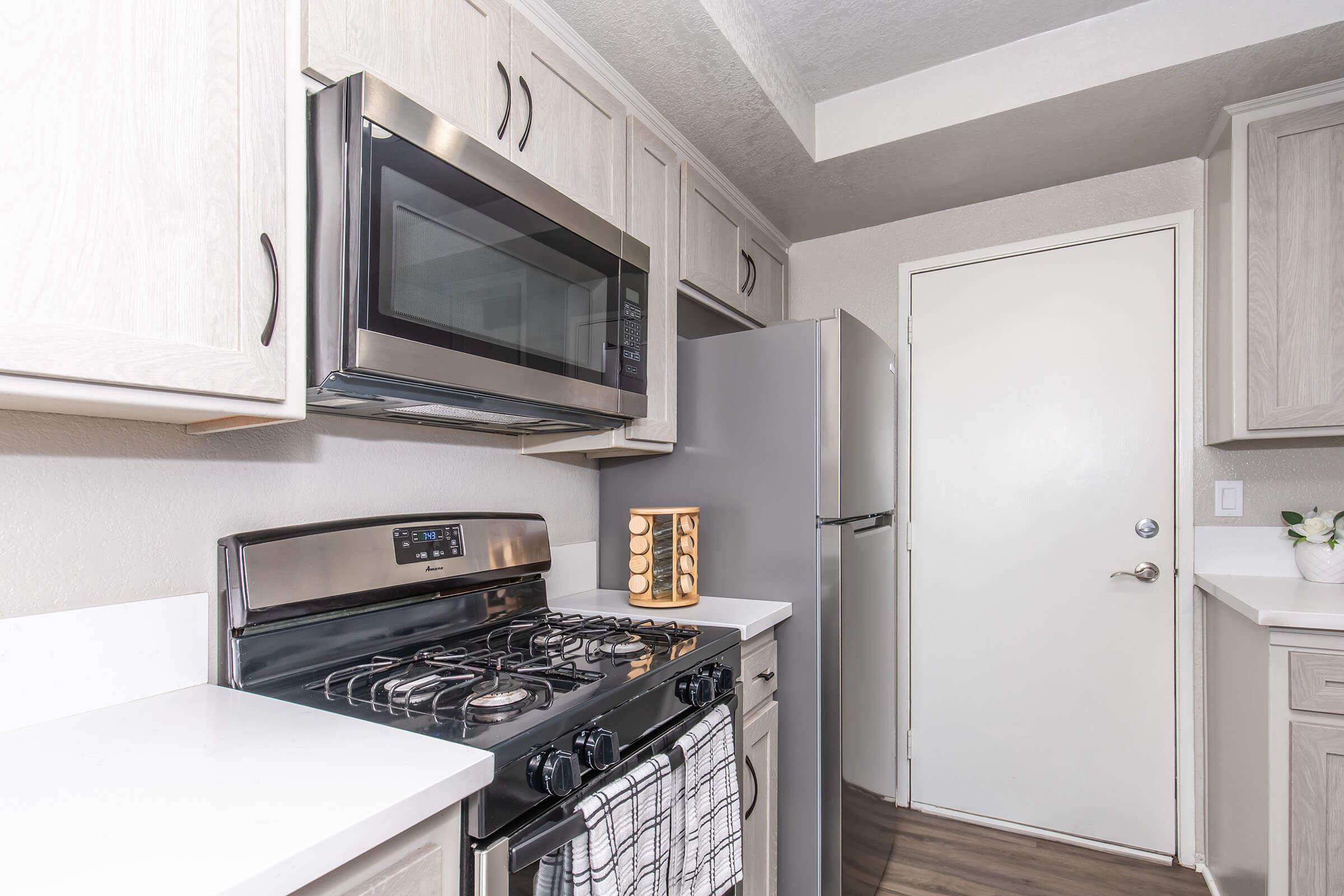 Modern kitchen featuring light wood cabinetry, stainless steel microwave and gas stove, white countertops, and a sleek refrigerator. A door leads out of the kitchen, and a decorative wooden item is placed on the counter. The flooring is a neutral tone.