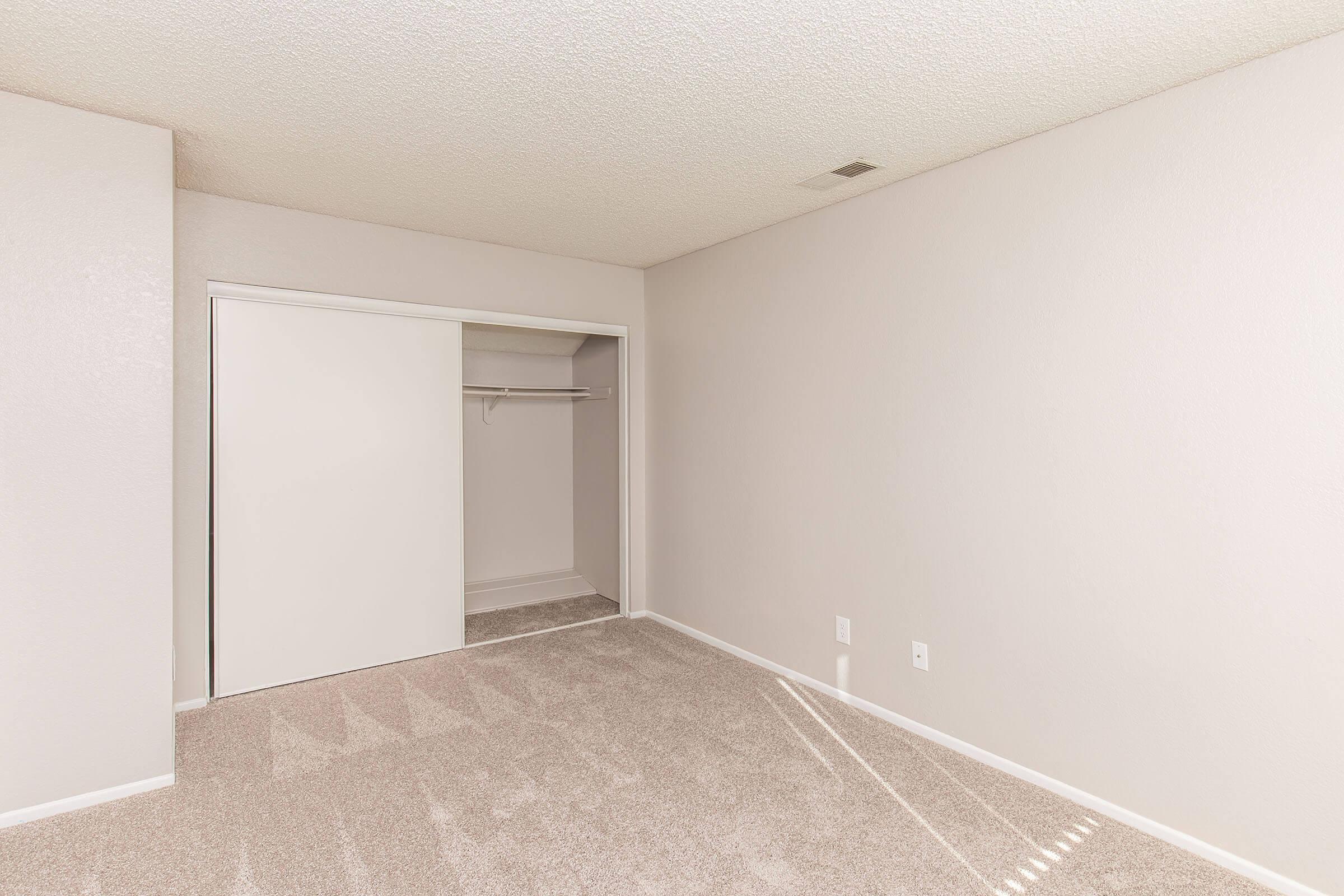 Empty, unfurnished room with light gray walls and carpet. A sliding door closet is seen on the left. The room has natural light coming in from a window, creating bright, shadowed patterns on the floor. The overall atmosphere is clean and minimalistic.