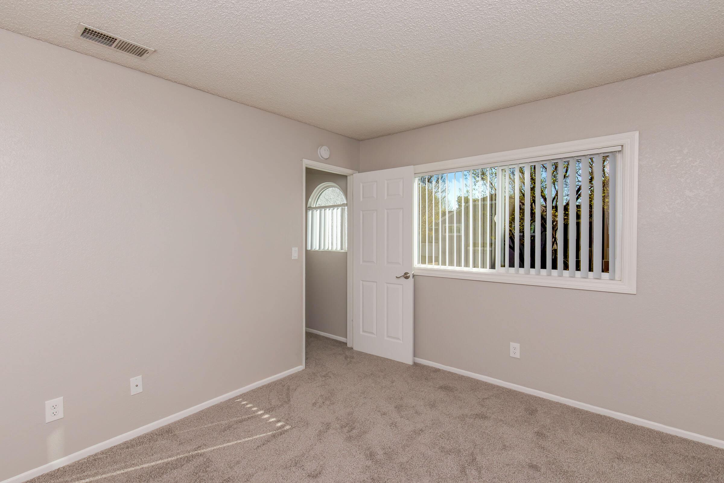 A well-lit, empty room with light gray walls and carpet. A window with white blinds lets in sunlight, illuminating the space. A doorway on the left leads to another area, and the overall atmosphere is clean and inviting, suitable for various uses such as a bedroom or office.