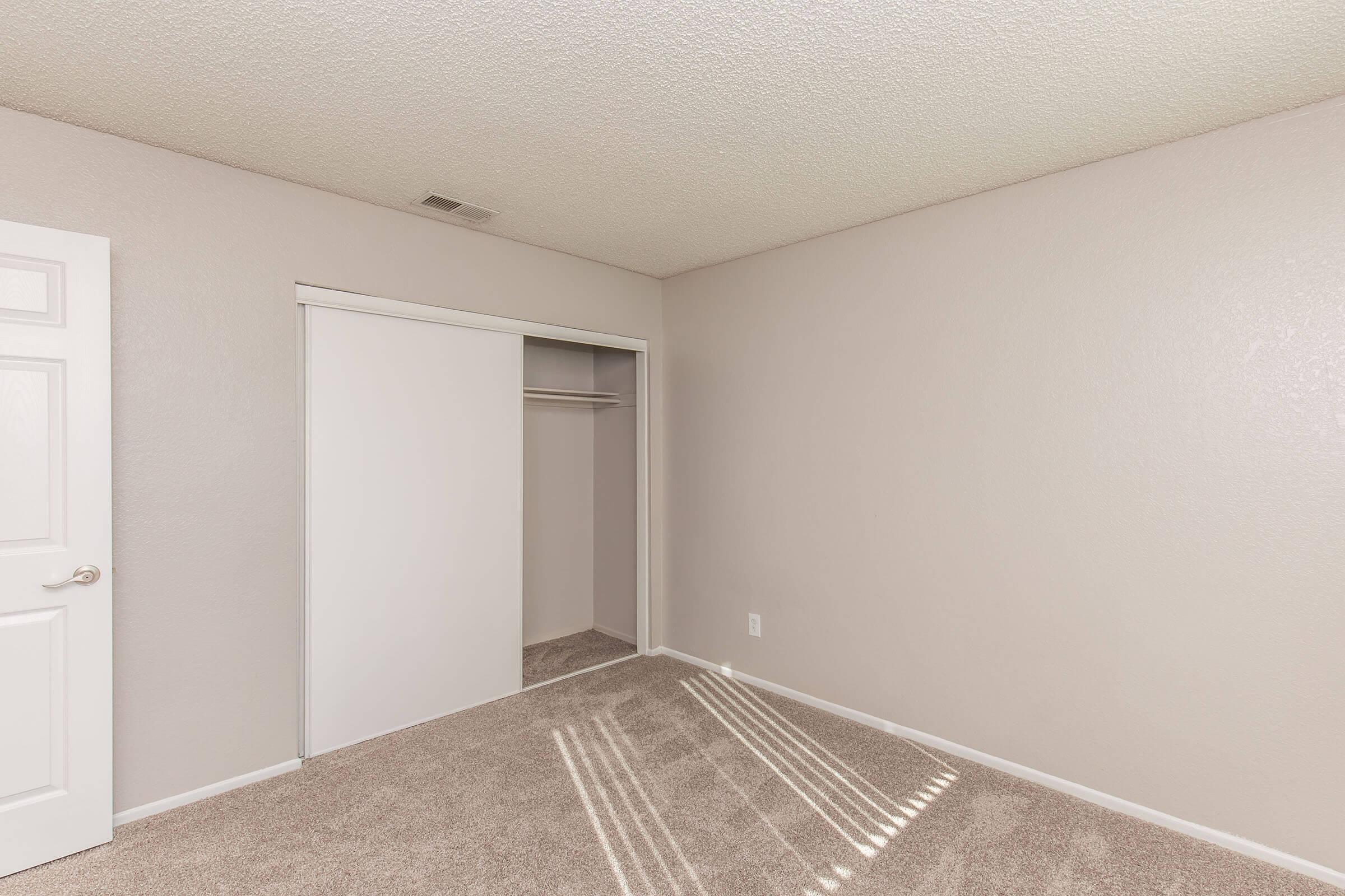 Empty room with light beige walls and carpeted floor. A white door is on the left, and there is a sliding closet door on the right. Soft shadows from sunlight create lines on the floor. The space appears clean and well-maintained, suggesting it could be used for various purposes.