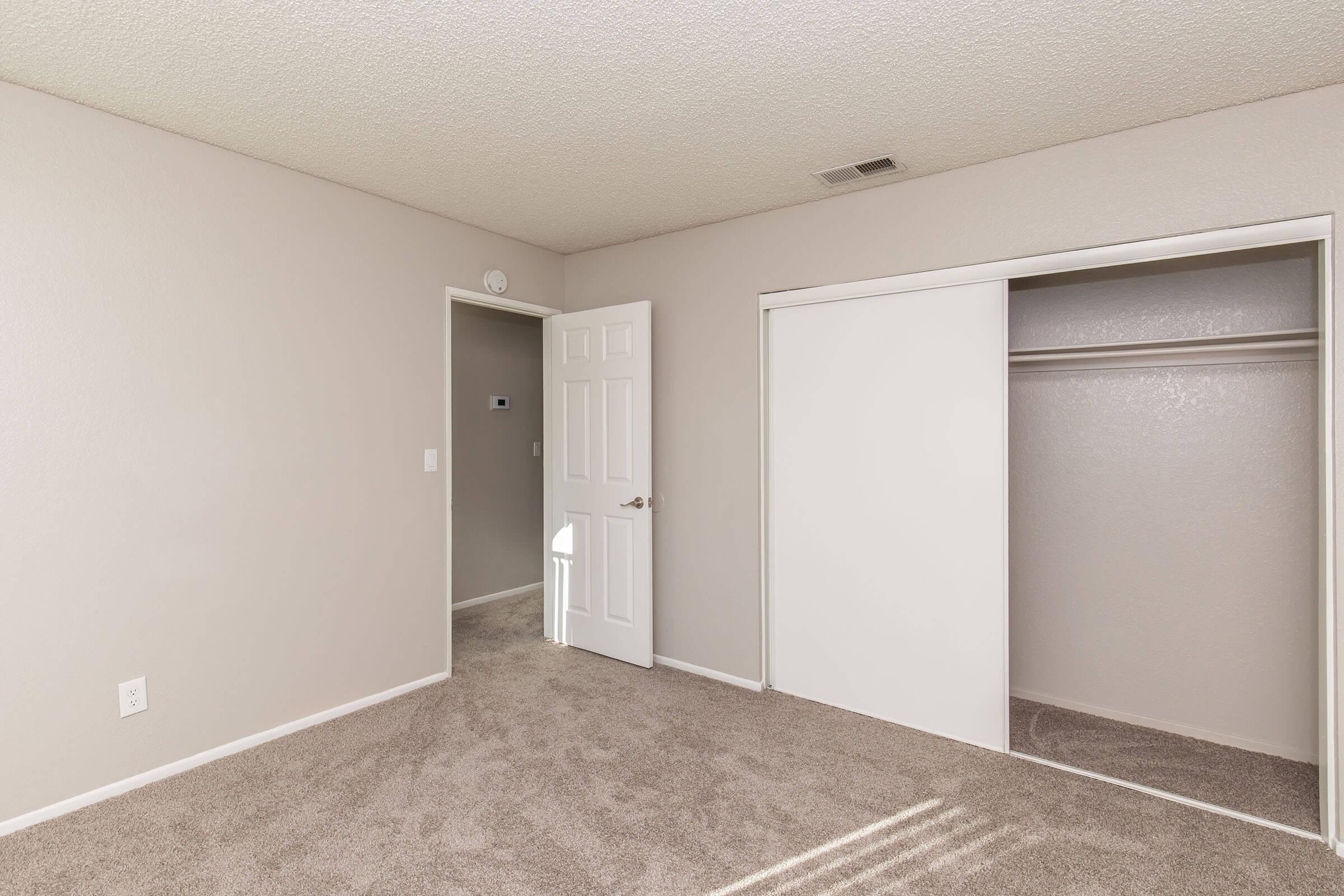 A clean, empty bedroom with beige walls and light carpet. There is a brown closet with sliding doors on one side, and a white door leading to another room. The space is unobstructed, indicating it's ready for moving in or decoration.