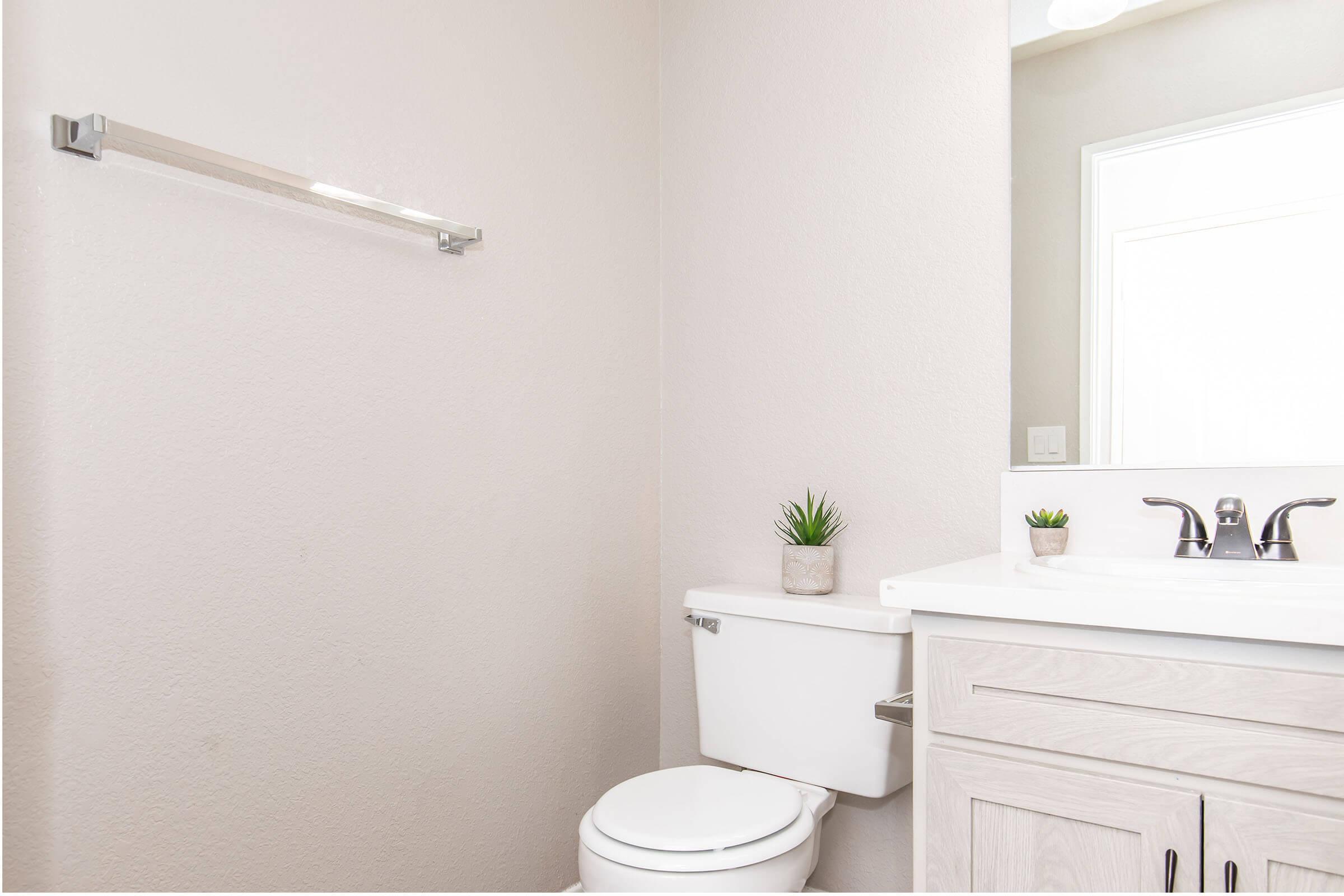 A clean, modern bathroom featuring a white toilet, a light-colored cabinet with a sink, and a mirror above the sink. There are small decorative plants on the countertop and a towel rack mounted on the wall. The walls are painted in a soft neutral color, creating a bright and airy atmosphere.