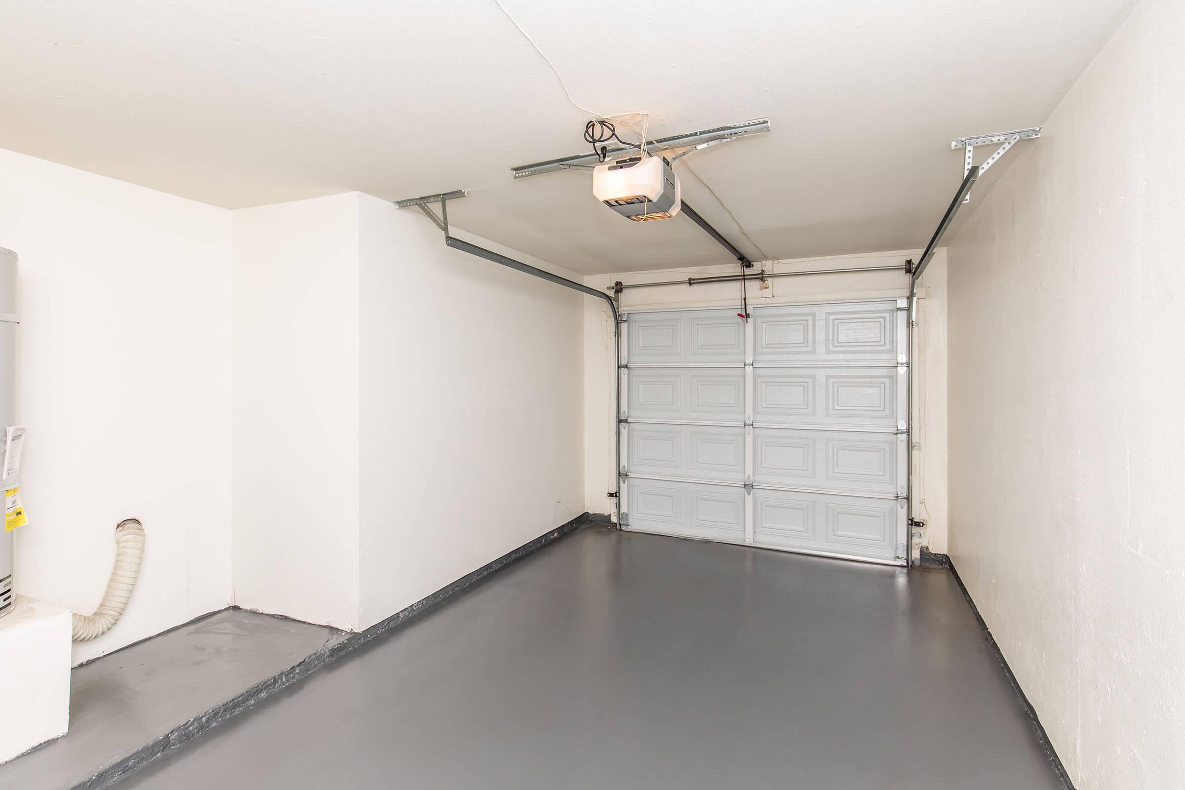 Empty garage interior featuring a grey painted floor, a white wall, and a garage door. The space is well-lit, with a ceiling-mounted garage door opener visible. A side wall includes a utility item. The overall ambiance is clean and spacious, suitable for vehicle storage or organization purposes.