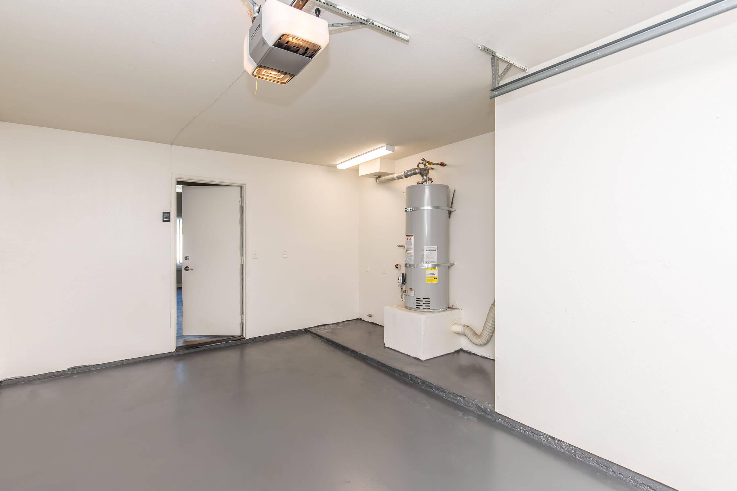 A clean and empty garage space with a gray floor, a water heater in the corner, and a door leading outside. The walls are white, and there is overhead lighting. A garage door opener is visible on the ceiling.