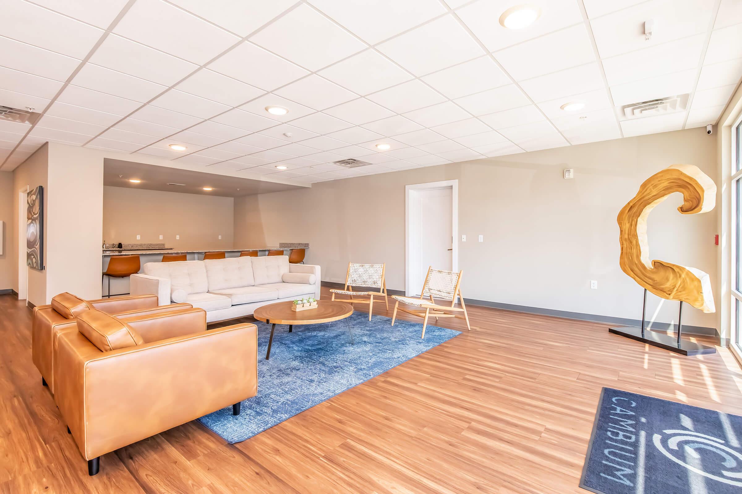 A spacious, modern lounge area featuring a light-colored sectional sofa, two wooden chairs, a round coffee table, and a large area rug. Natural light floods the room through large windows, highlighting the wooden accents and minimalist decor. A stylish wooden sculpture and a welcome mat add character to the space.