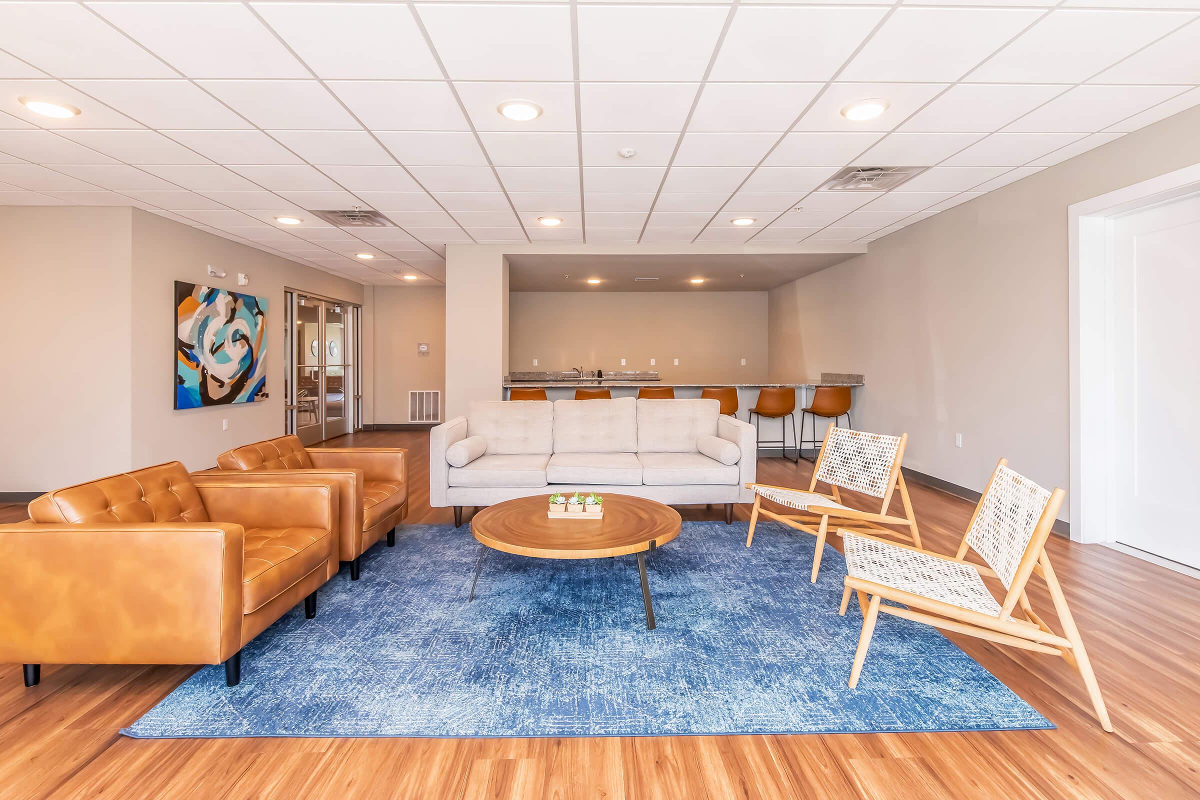 A modern living room featuring a light beige sofa, two brown leather chairs, and a round wooden coffee table on a blue area rug. There are three stylish wooden chairs with woven seats, and a bar area with high stools in the background, along with an abstract painting on the wall. Bright, open space with wooden floors.